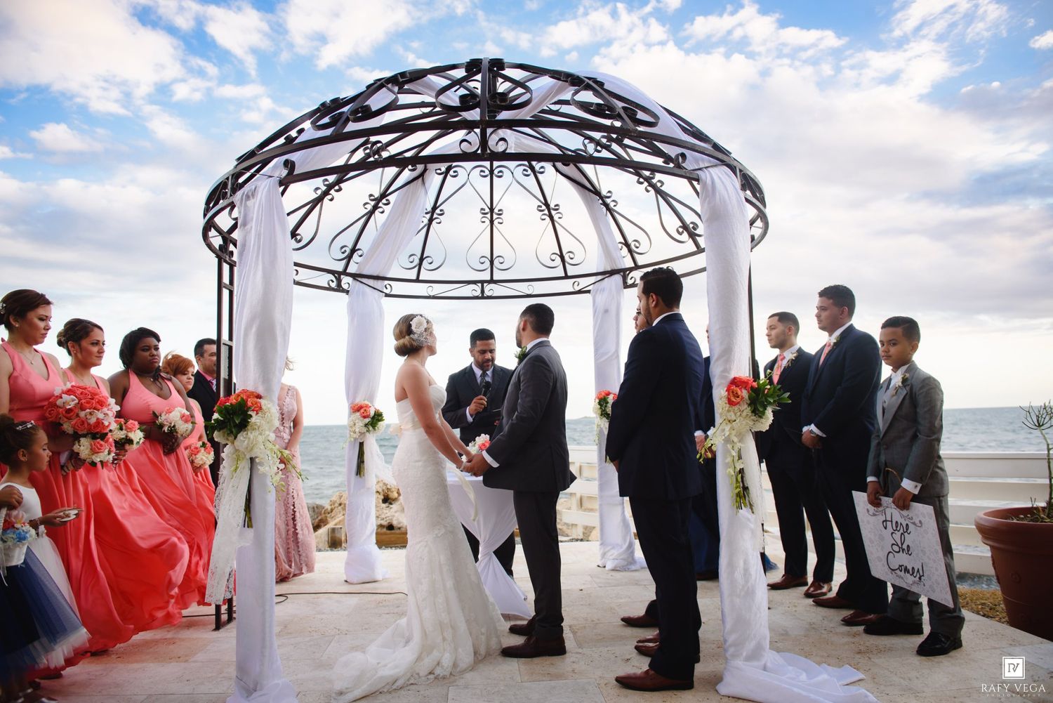 Outdoor beach wedding ceremony under ornate iron gazebo with bride, groom, officiant, bridesmaids in coral dresses, and groomsmen.