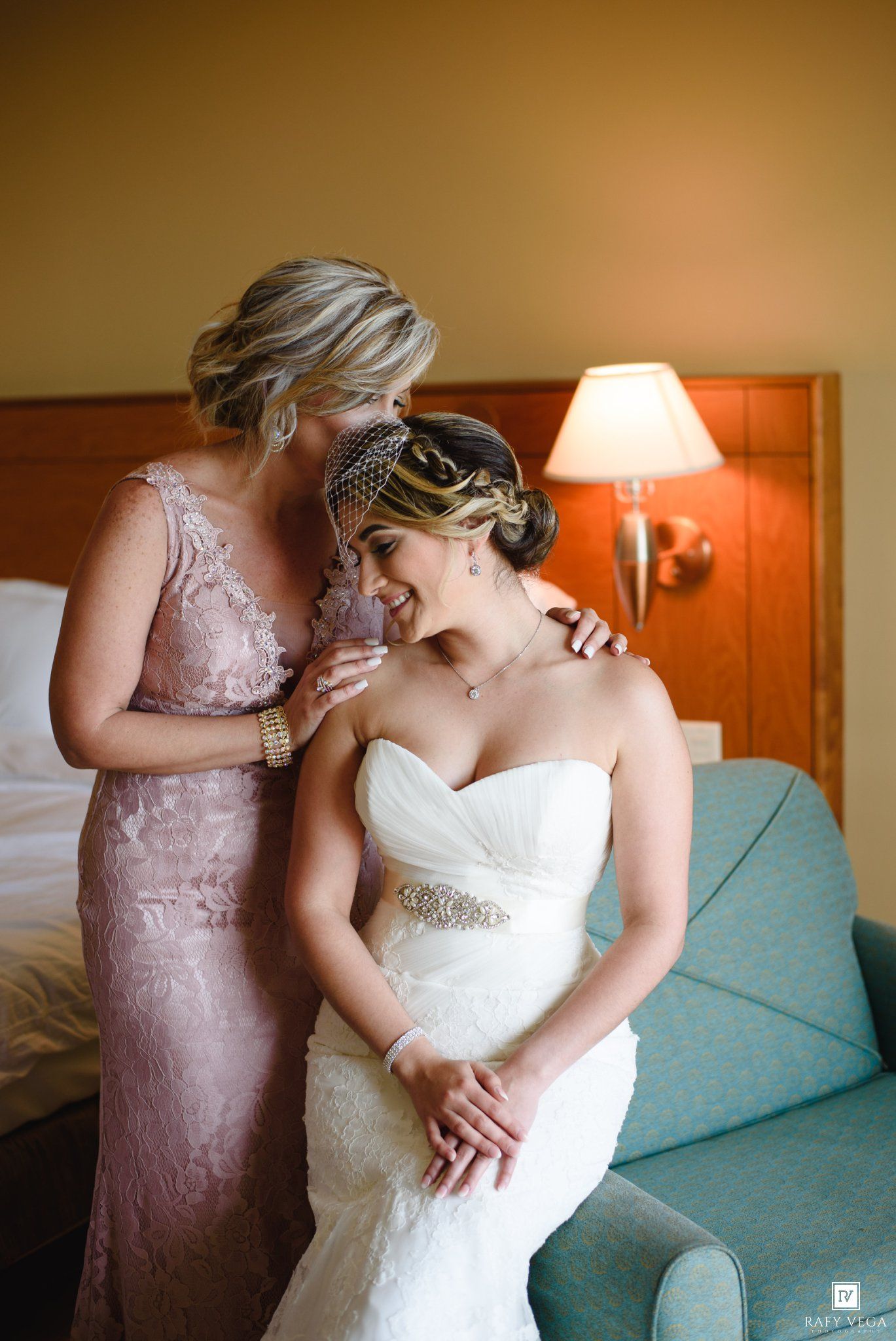 Bride in white strapless gown sits as woman in pink lace dress whispers and embraces her in hotel room.