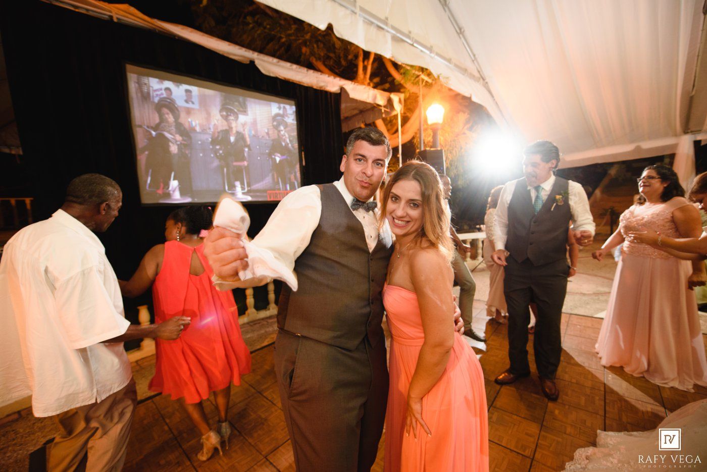 Couple smiling and posing at an elegant outdoor wedding reception with guests dancing under a white tent.