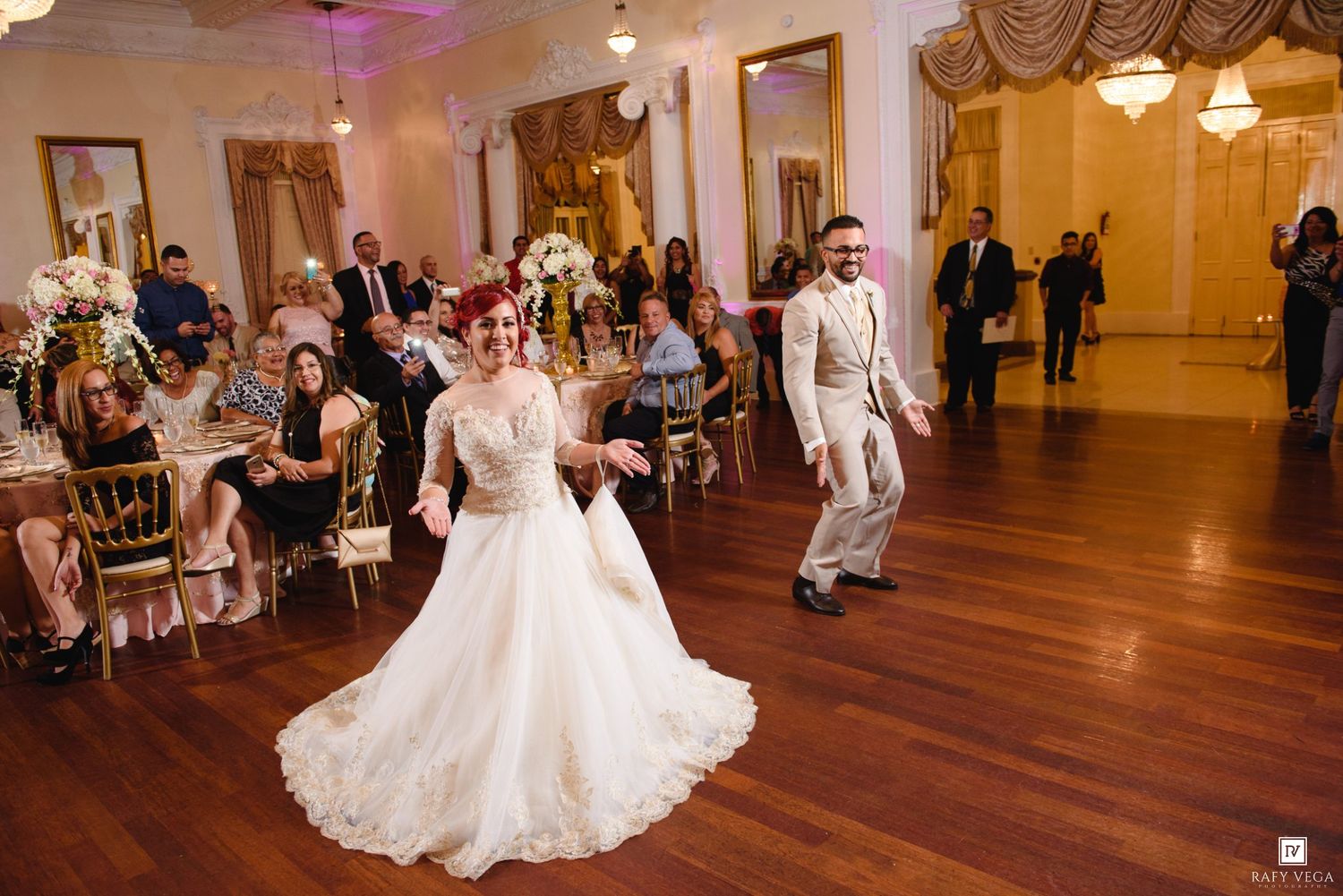 Bride in white gown and groom in ivory suit make grand entrance at elegant ballroom wedding reception.