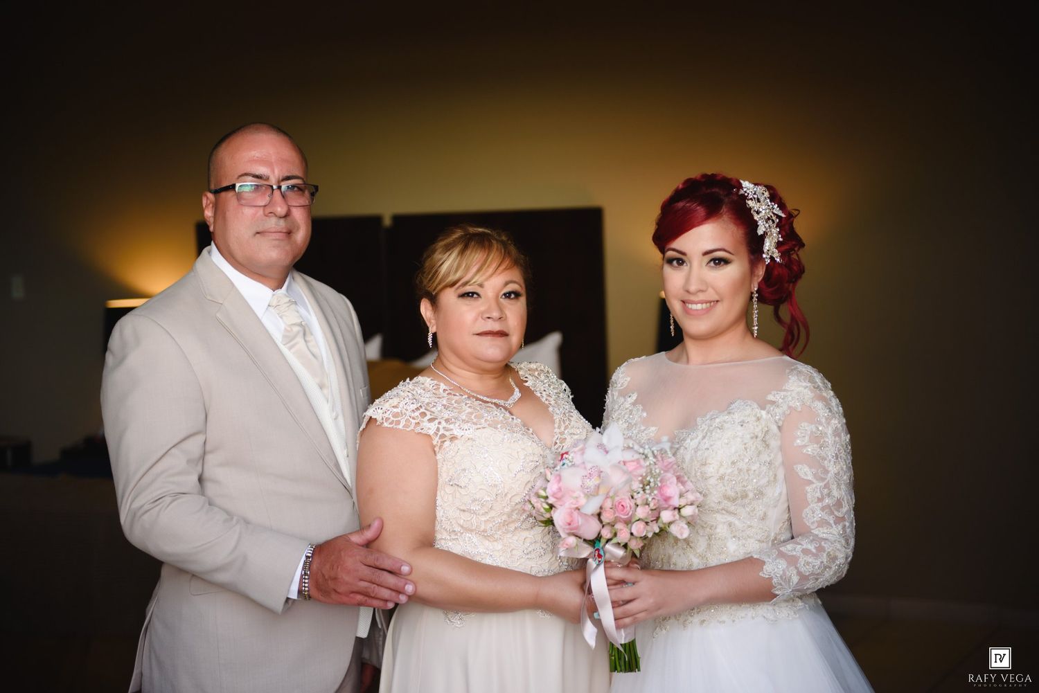 Bride in lace gown holding pink bouquet poses with two guests in elegant wedding venue photo.