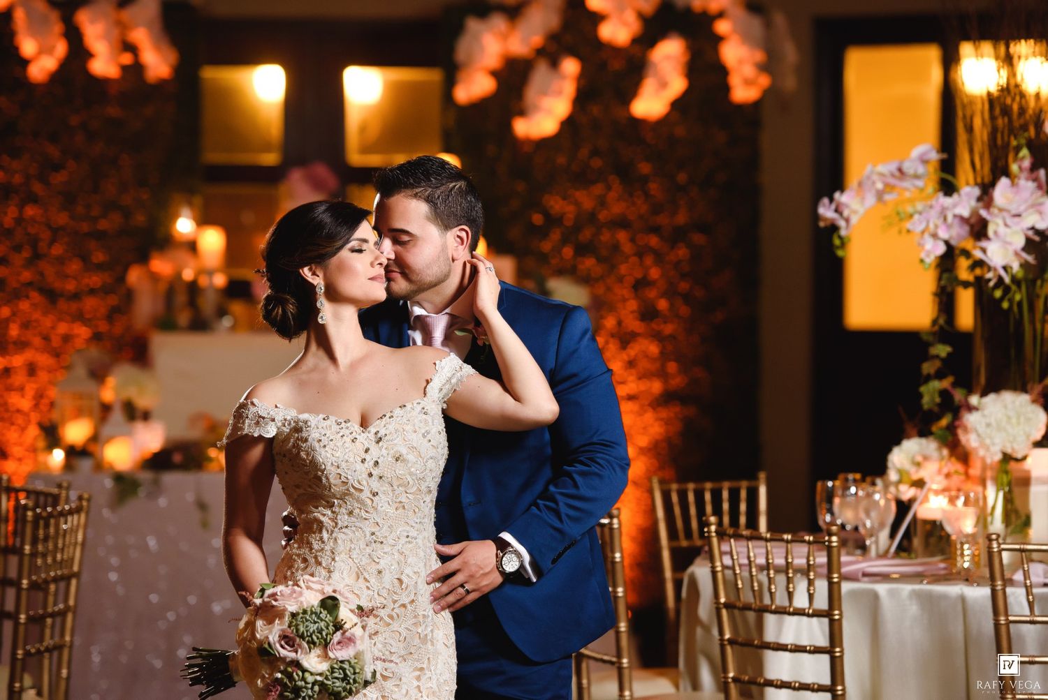 Bride and groom share romantic kiss in elegant wedding reception venue with warm golden lighting and floral decor.