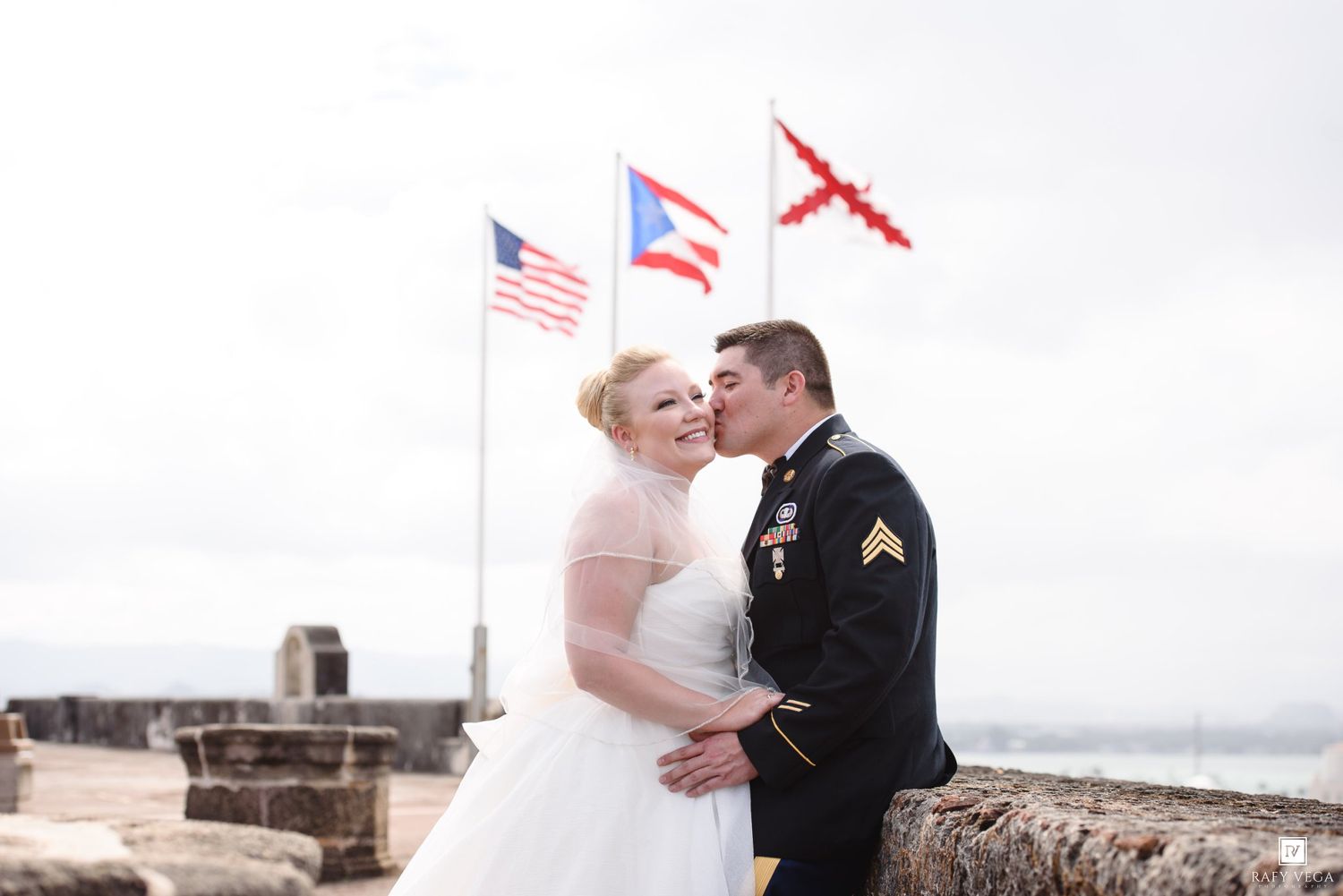 Military groom in uniform kisses bride in white dress at historic fort with American and Puerto Rican flags behind them.