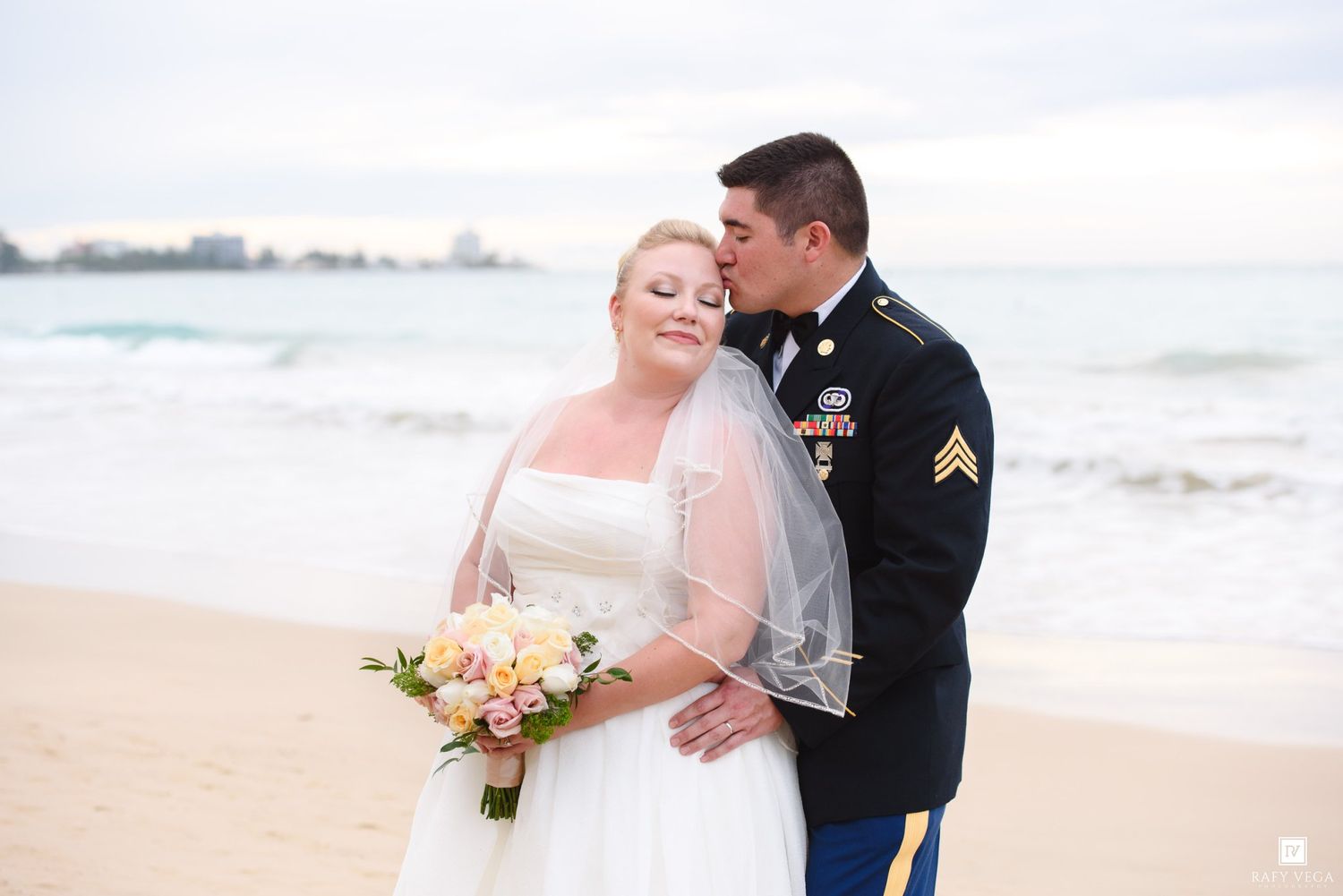 Military groom in dress uniform kissing bride in white gown on beach, bride holding pastel bouquet.
