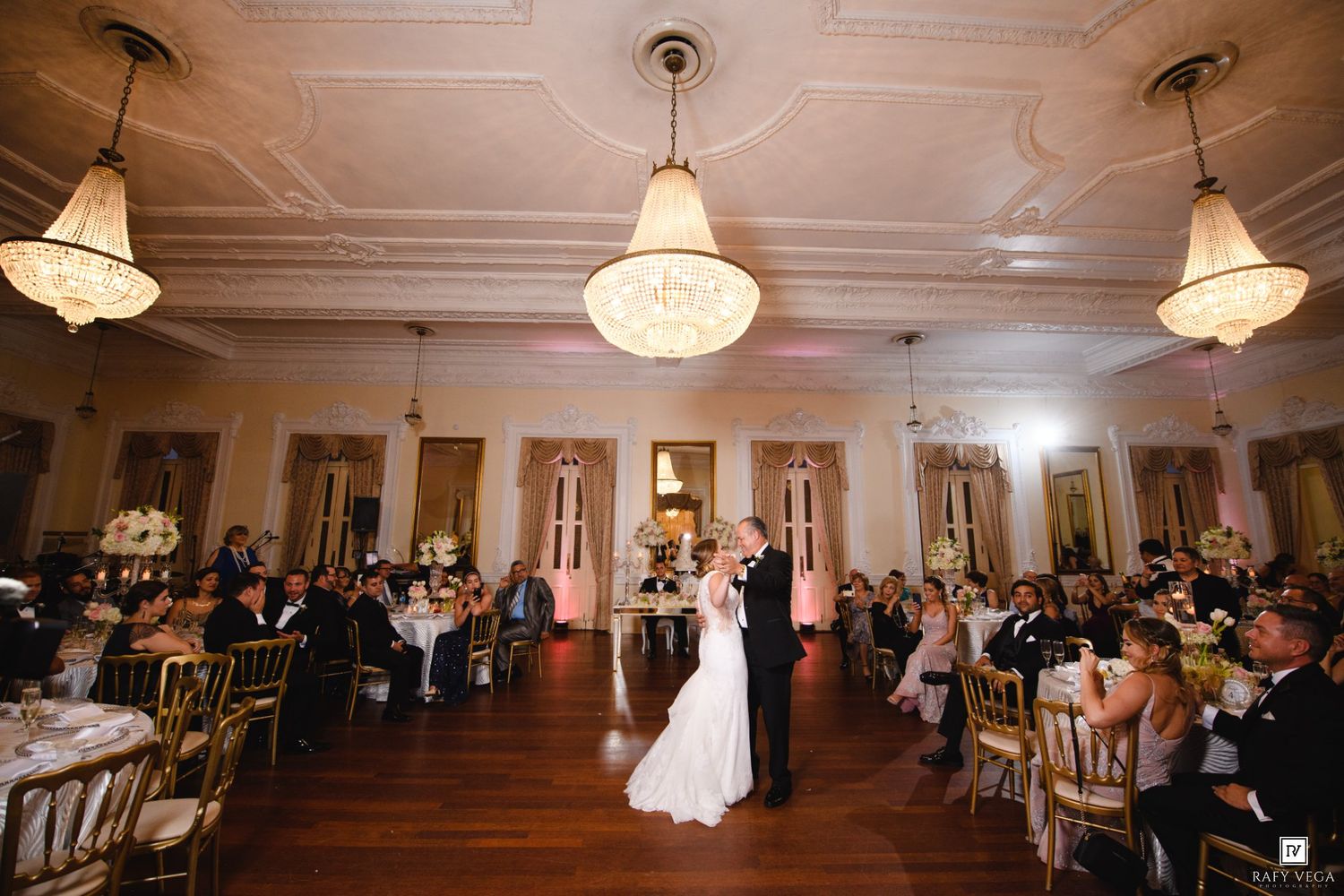 Bride and groom share first dance in elegant ballroom with chandeliers and guests seated at gold chairs.