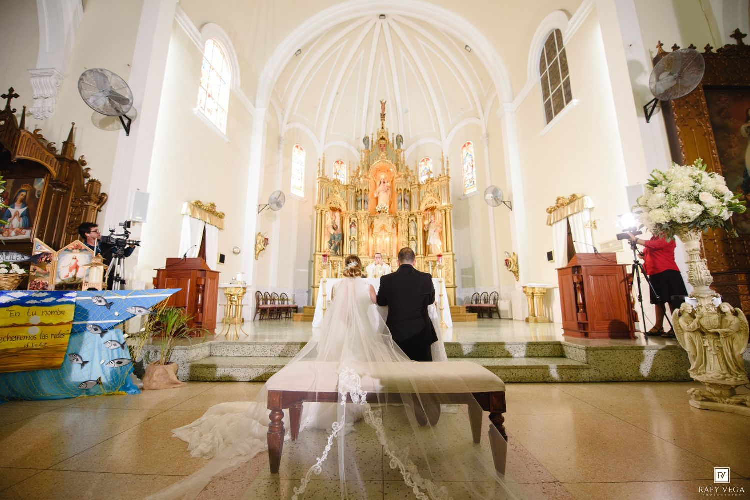 Bride and groom kneeling at the altar of an ornate white Catholic church during their wedding ceremony.