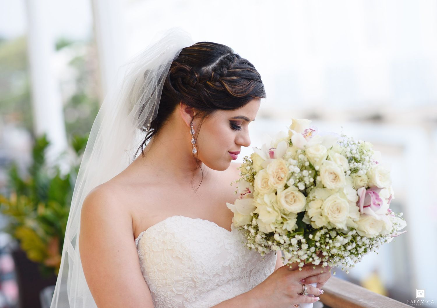 Elegant bride in strapless lace gown smelling white rose bouquet with braided updo and veil.