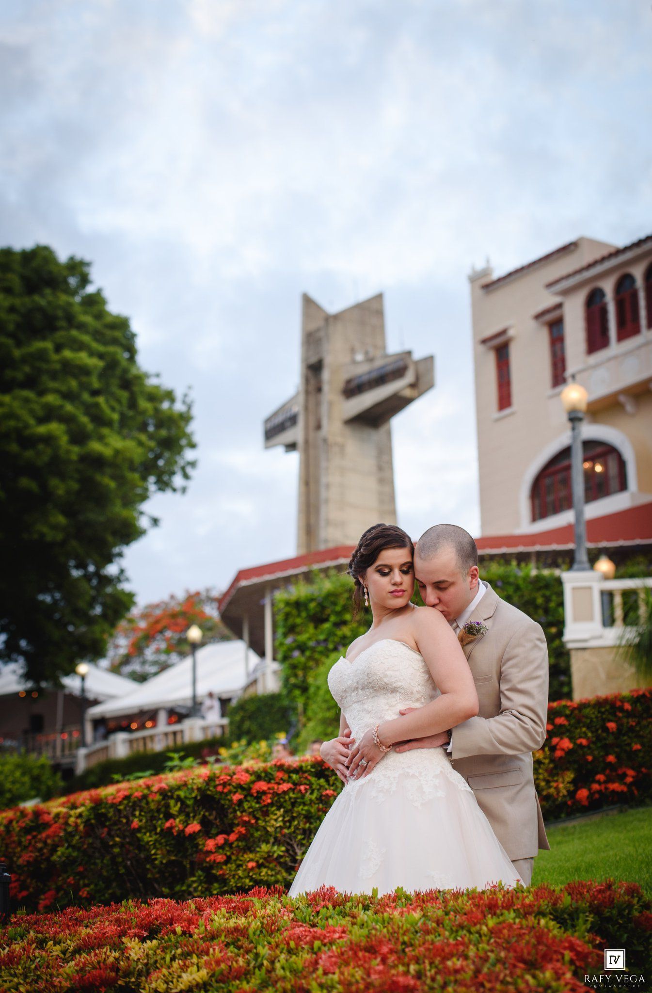 Bride and groom embrace in garden with floral blooms, historic church and large cross in background on their wedding day.