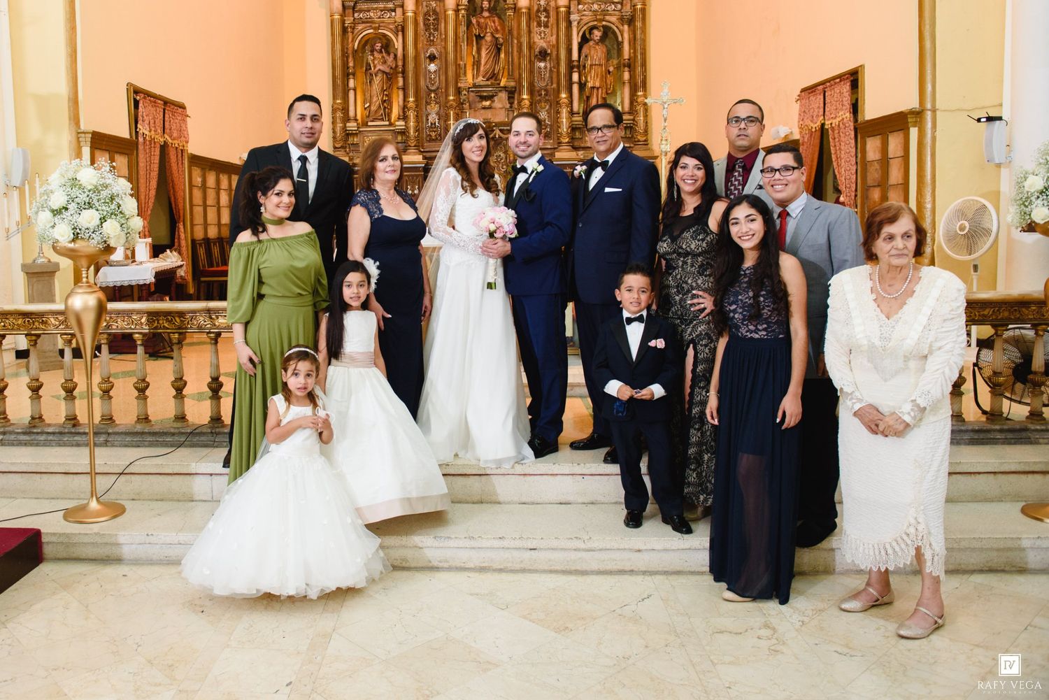 Wedding party poses on church altar steps, bride in white gown with groom and family including flower girls.