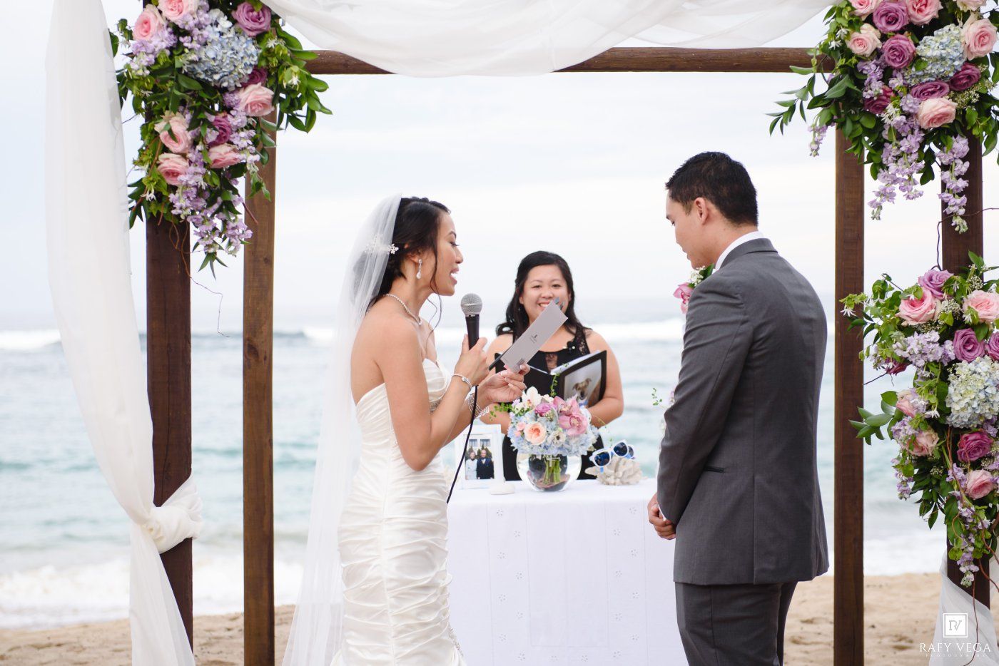 Bride reads vows to groom at romantic beach wedding ceremony under floral arch with ocean backdrop.