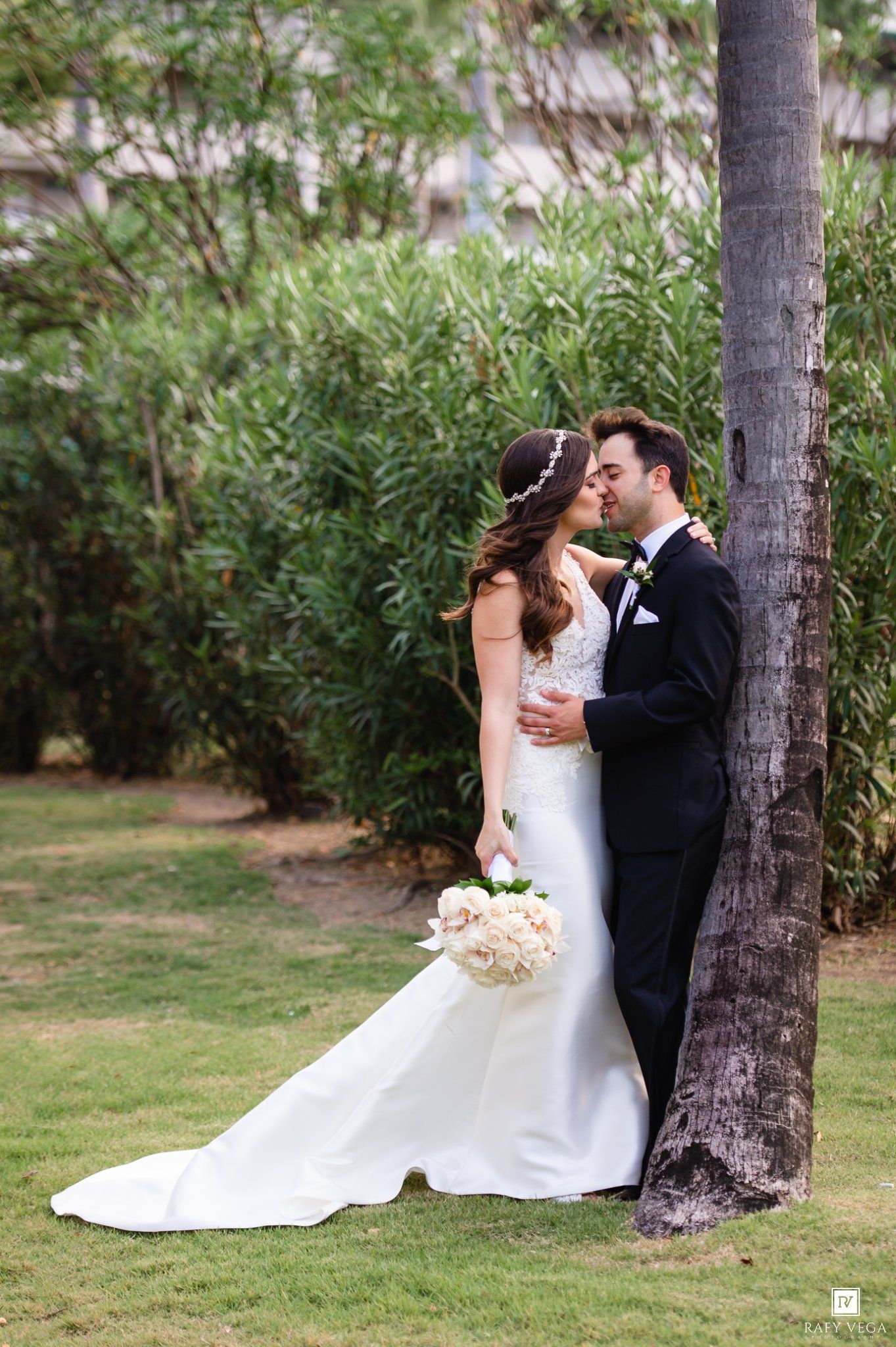 Bride and groom sharing a romantic kiss leaning against a palm tree outdoors.