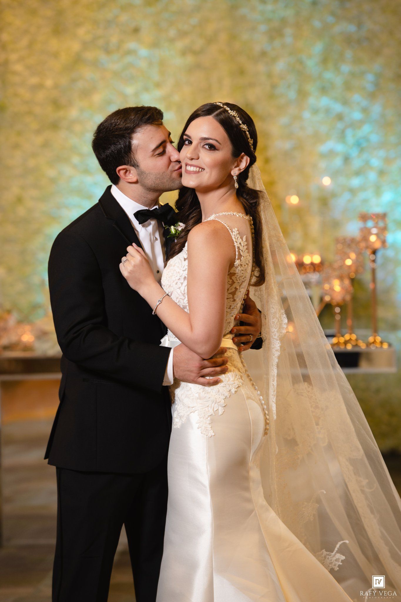 Bride in white lace gown with veil and groom in black tuxedo share a romantic kiss at elegant wedding reception.