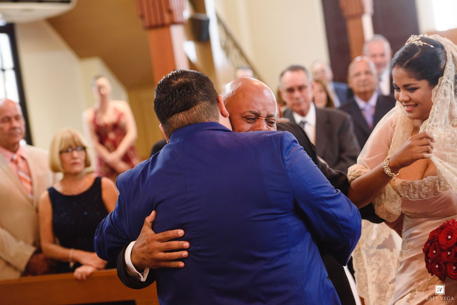 Groom in blue suit embracing a guest at a wedding ceremony while bride in white lace dress watches nearby.