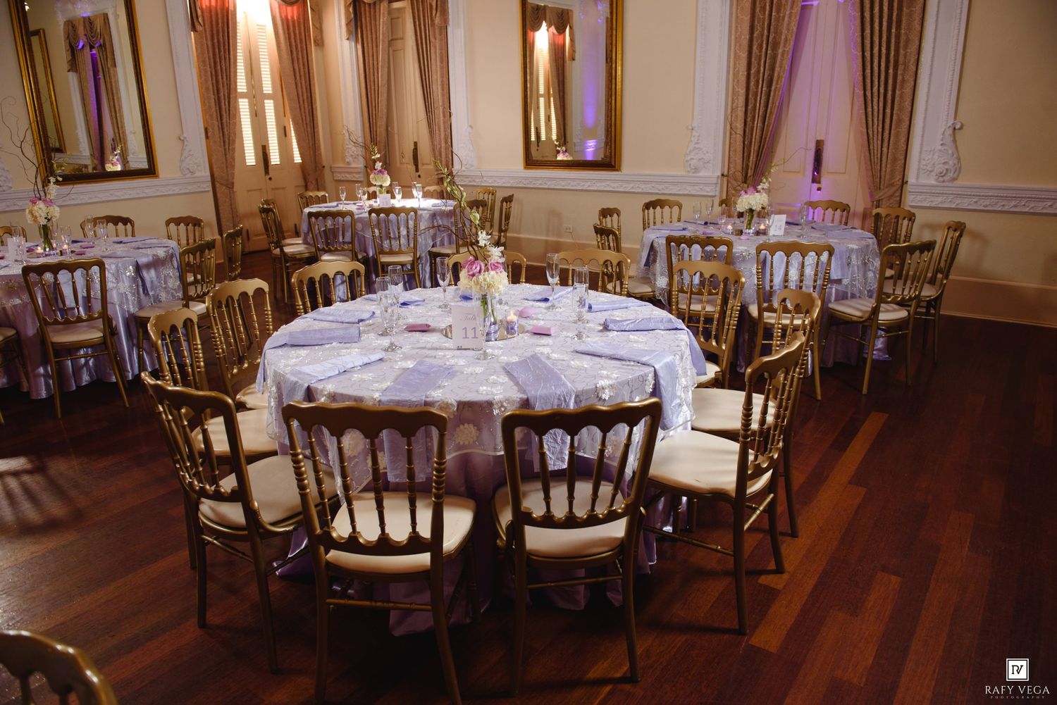 Elegant banquet hall with round tables, white linens, and gold Chiavari chairs on hardwood floors.