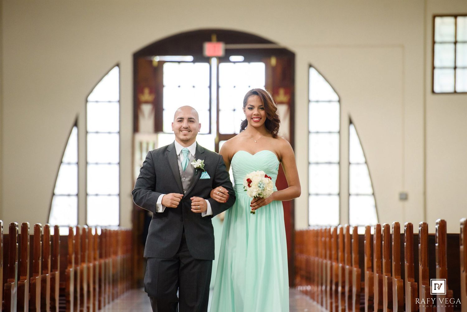 Bridesmaid in mint green gown walks down church aisle with groomsman in gray suit during wedding ceremony.