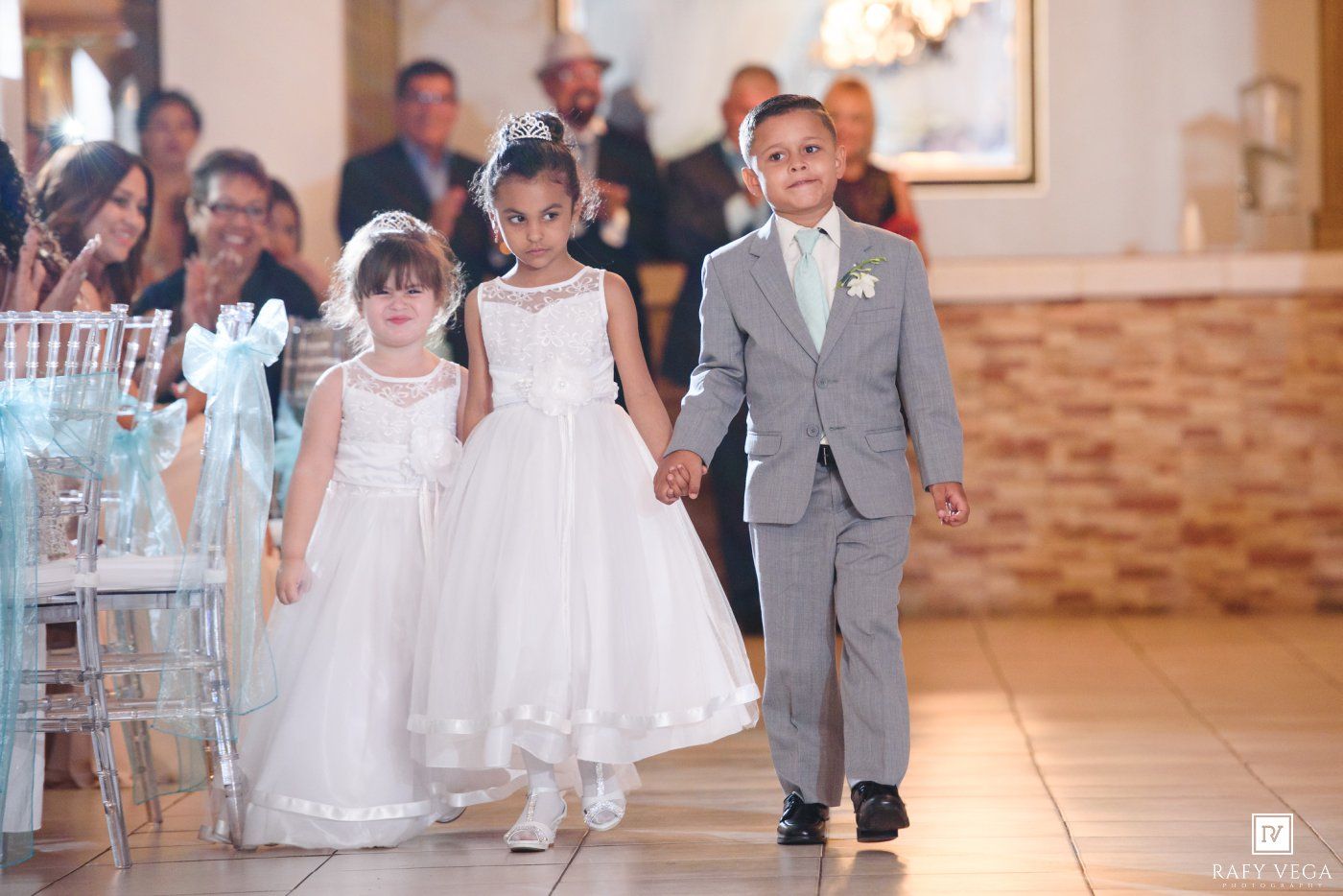 Three children in formal wedding attire walk down the aisle, two flower girls in white dresses and a ring bearer in a gray suit.
