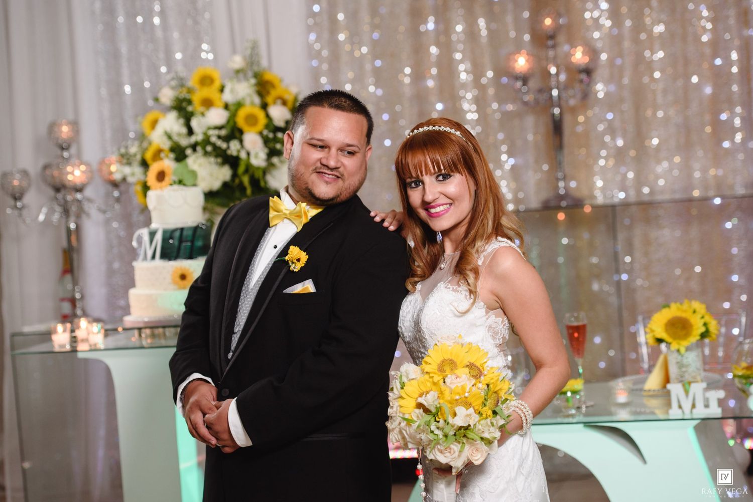 Newlyweds posing together at wedding reception with sunflower bouquet and decorated cake table behind them.