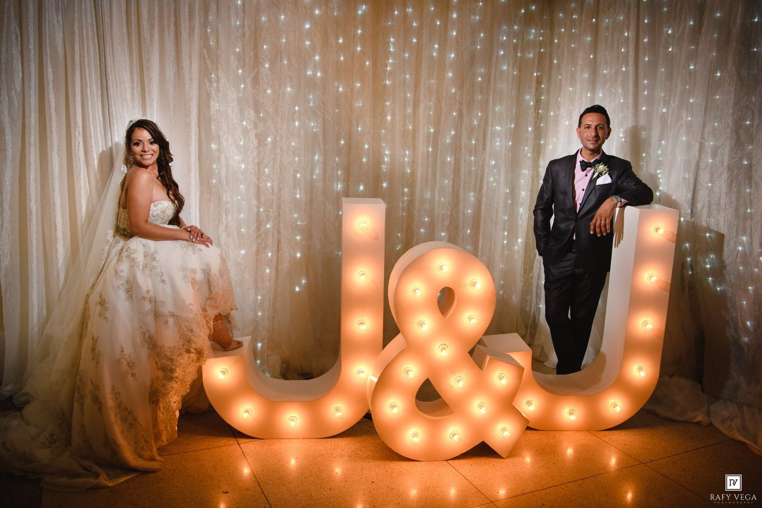 Bride and groom posing beside illuminated marquee letters J&U at elegant wedding reception with twinkling backdrop.