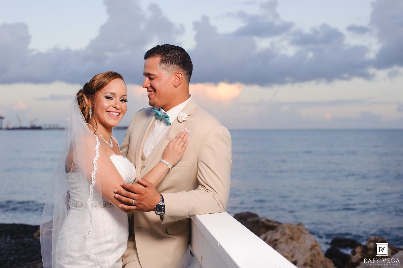 A bride and groom share a romantic moment by the ocean at sunset, with the groom in a beige suit and teal bow tie.