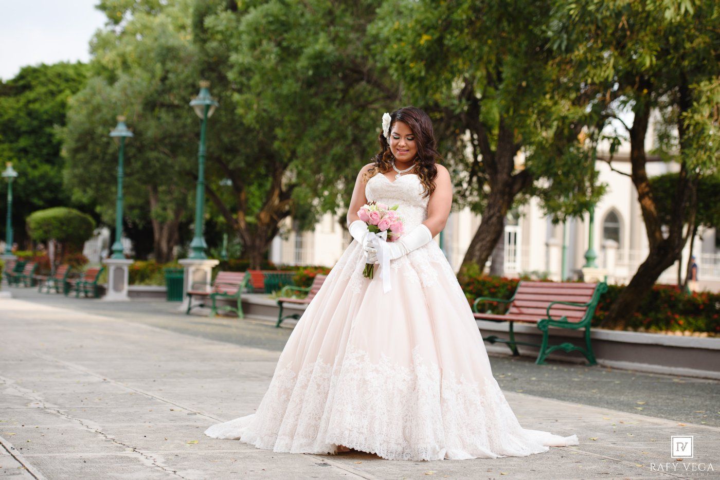 Bride in white ball gown holding pink bouquet poses outdoors near park benches and trees on a sunny day.