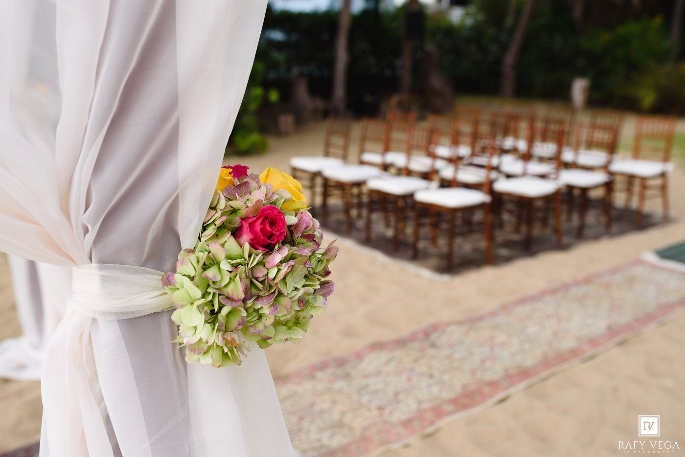 Outdoor wedding ceremony setup with wooden chairs, a floral bouquet tied to a white draped curtain in the foreground.