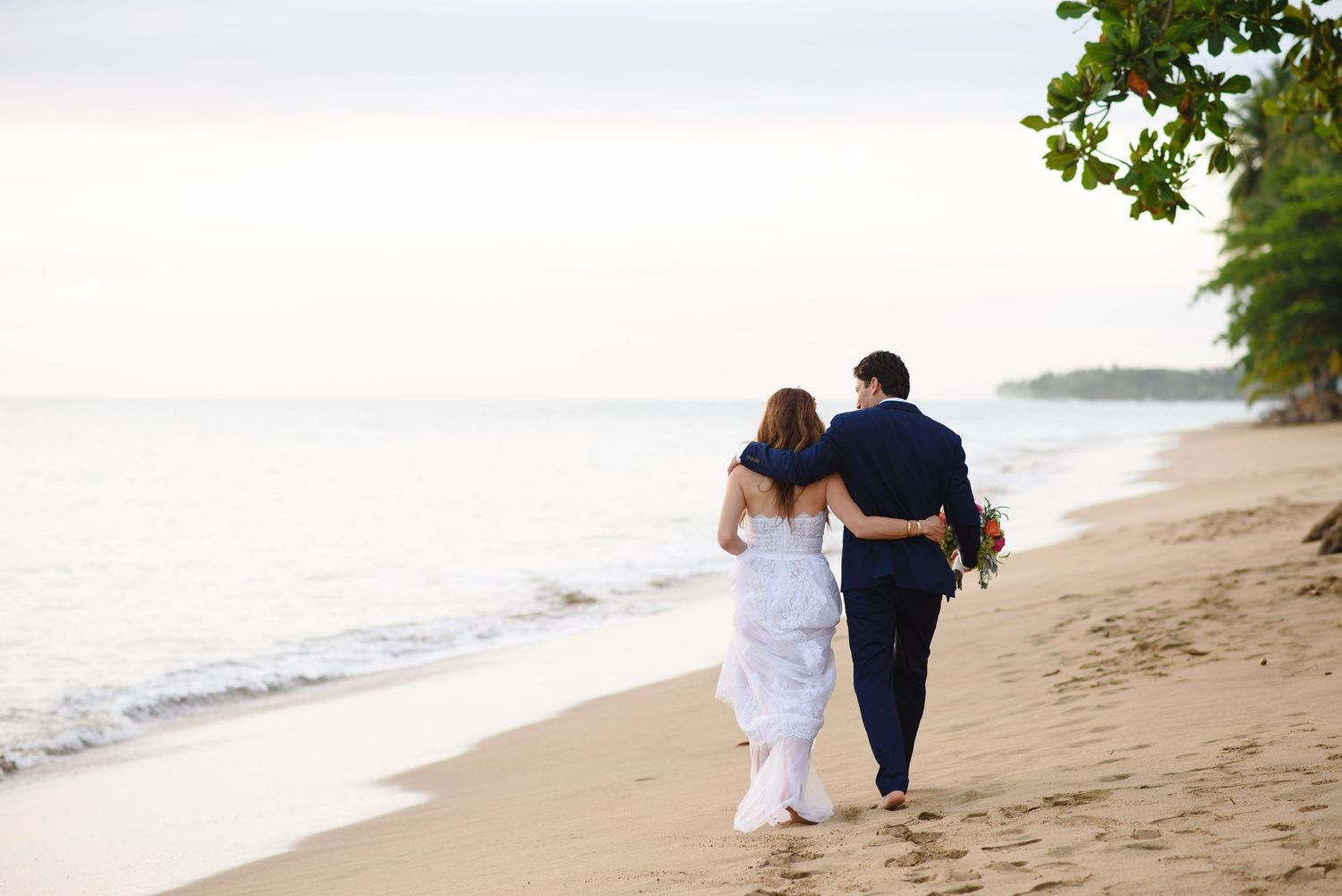 Newlywed couple walking hand in hand along a tropical beach at sunset in wedding attire.