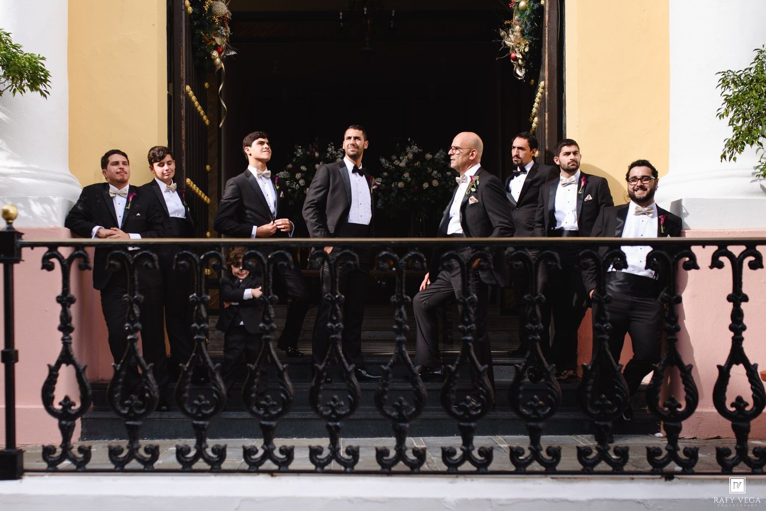 Groomsmen in black tuxedos posing on an ornate wrought iron balcony at a formal wedding venue.