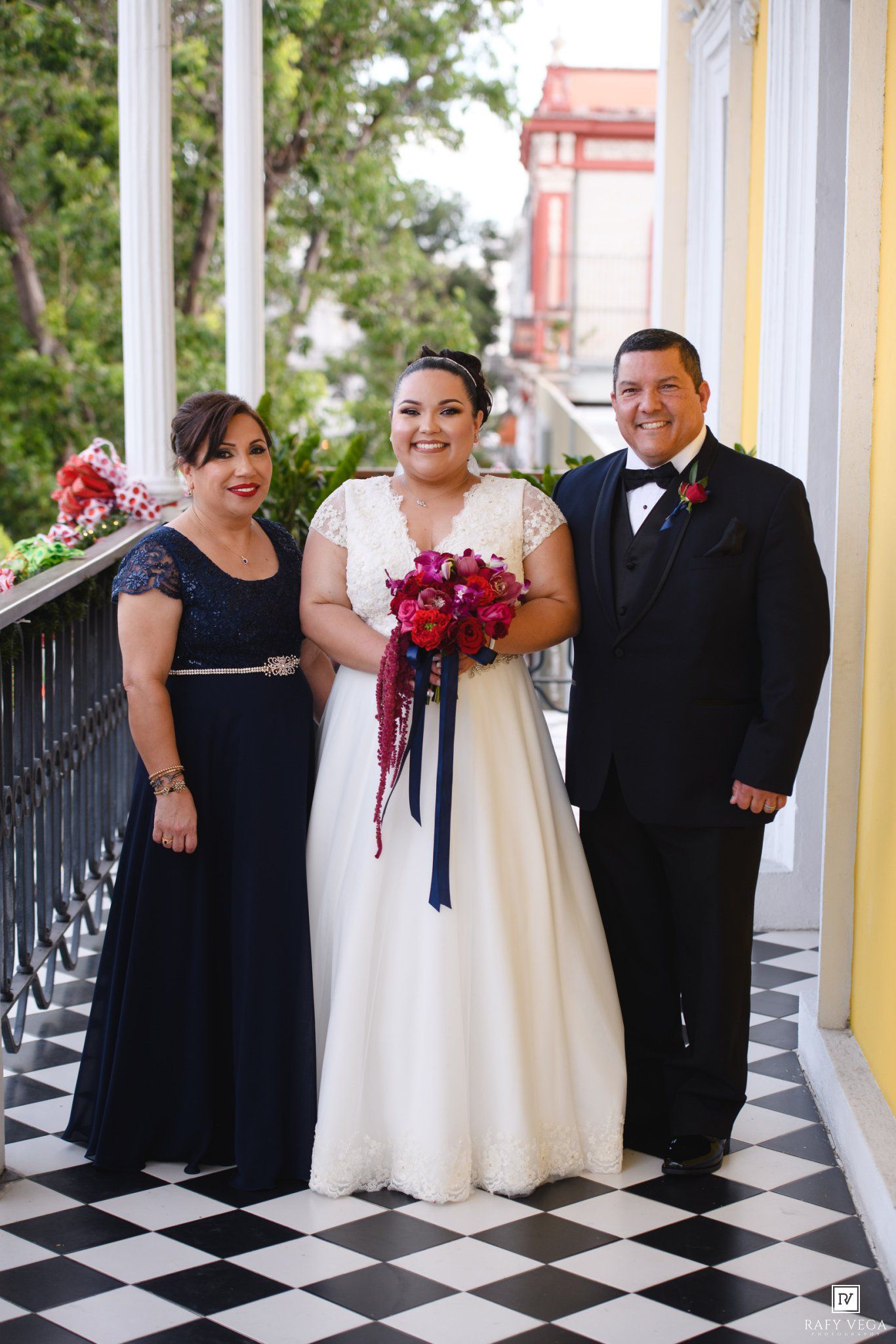 Bride in white gown holding red bouquet poses with parents on checkered floor balcony.