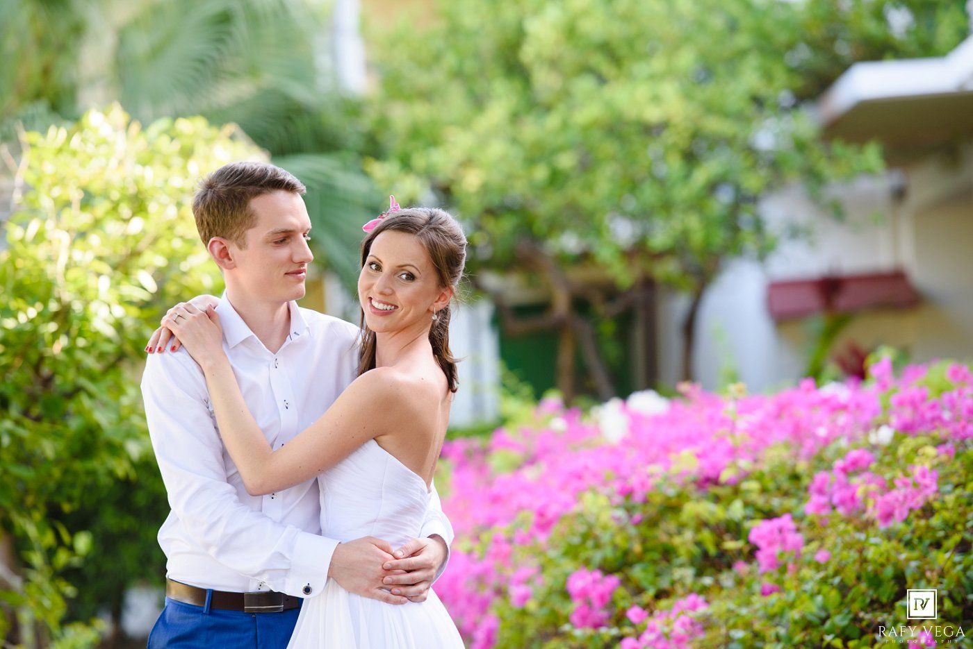 A happy couple embraces in a lush garden with vibrant pink flowers, dressed in white wedding attire.