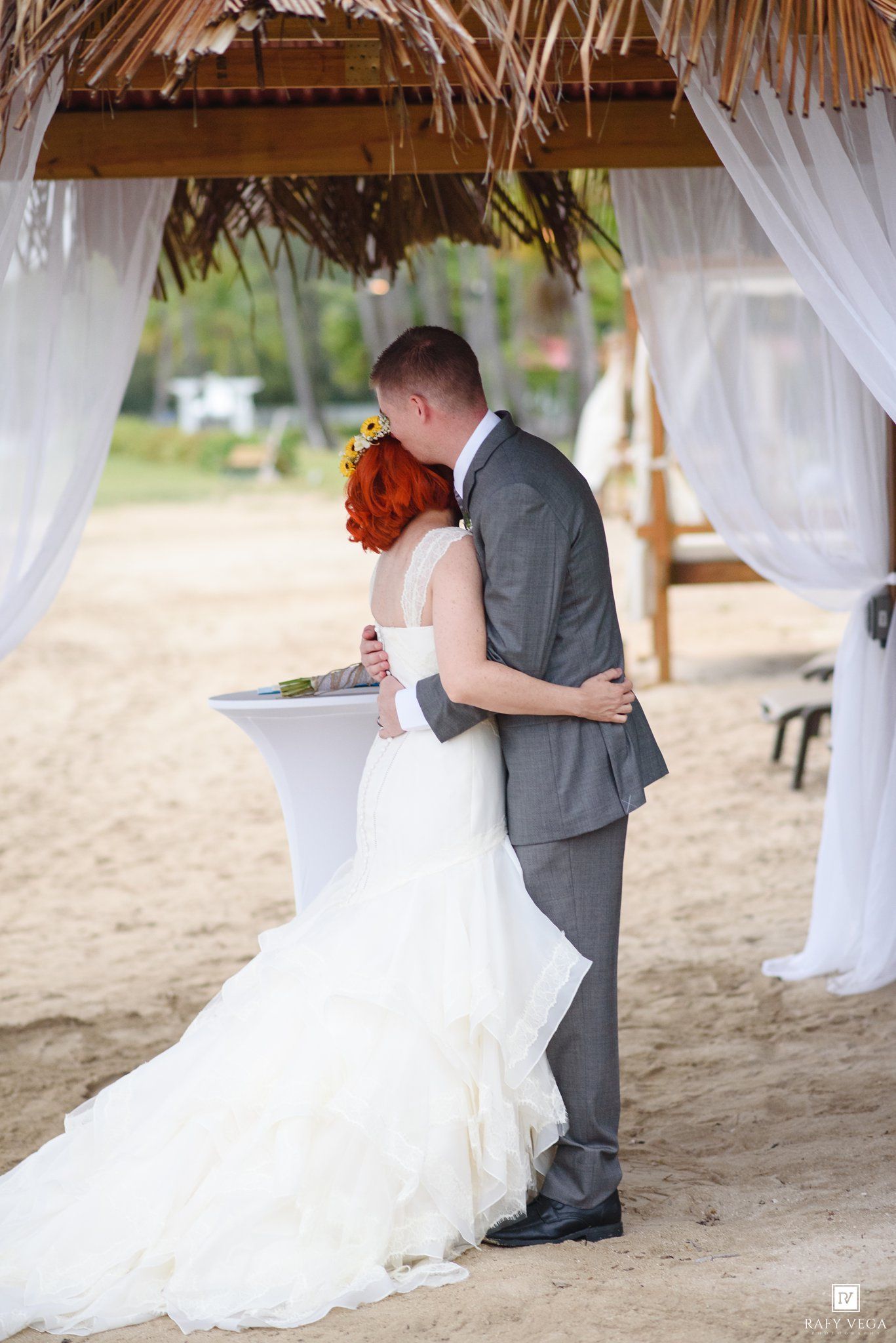 Bride in white ruffled gown and groom in gray suit share a kiss under a thatched beach gazebo with white drapes.