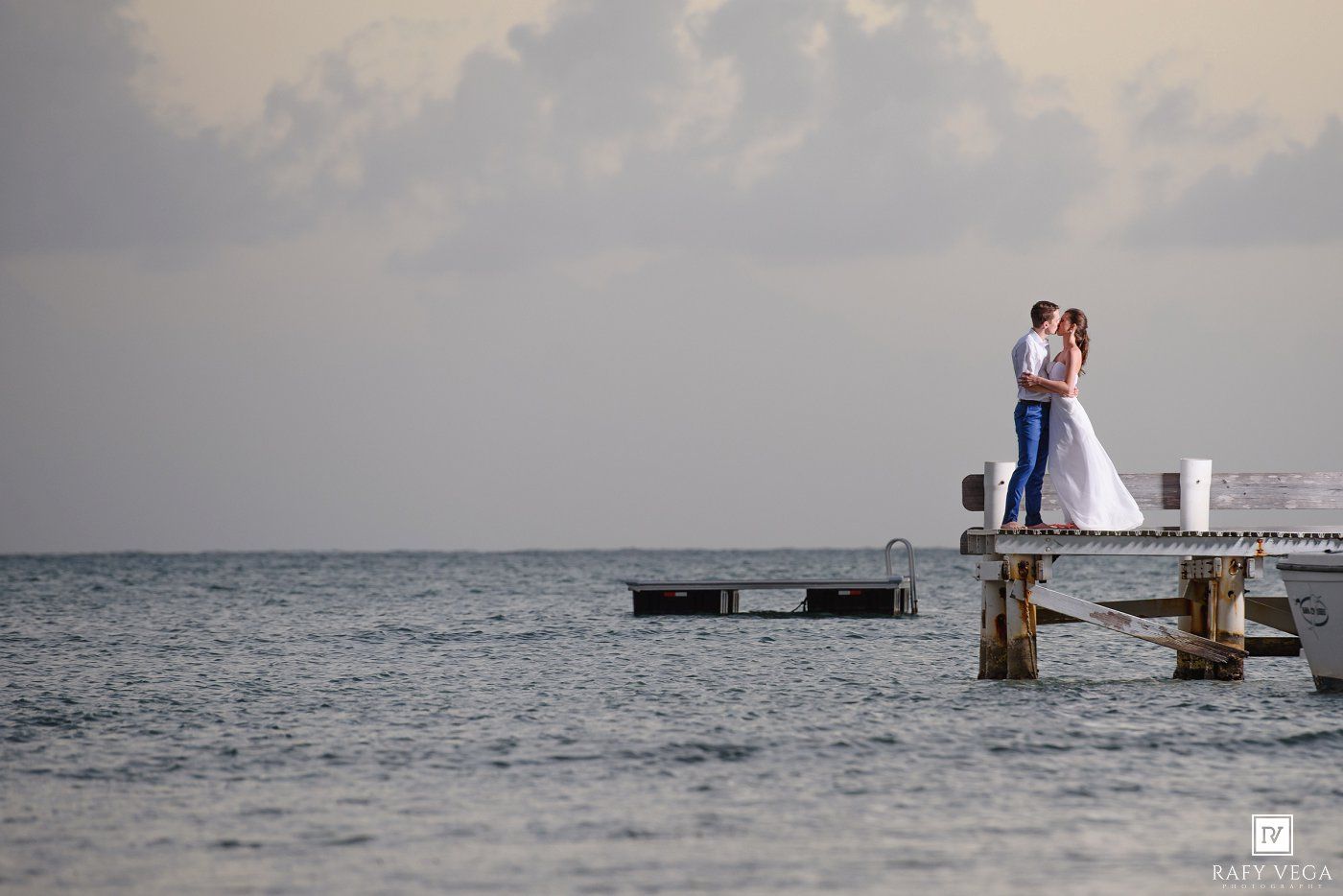Bride and groom share a romantic kiss on a wooden pier over calm ocean waters at dusk.