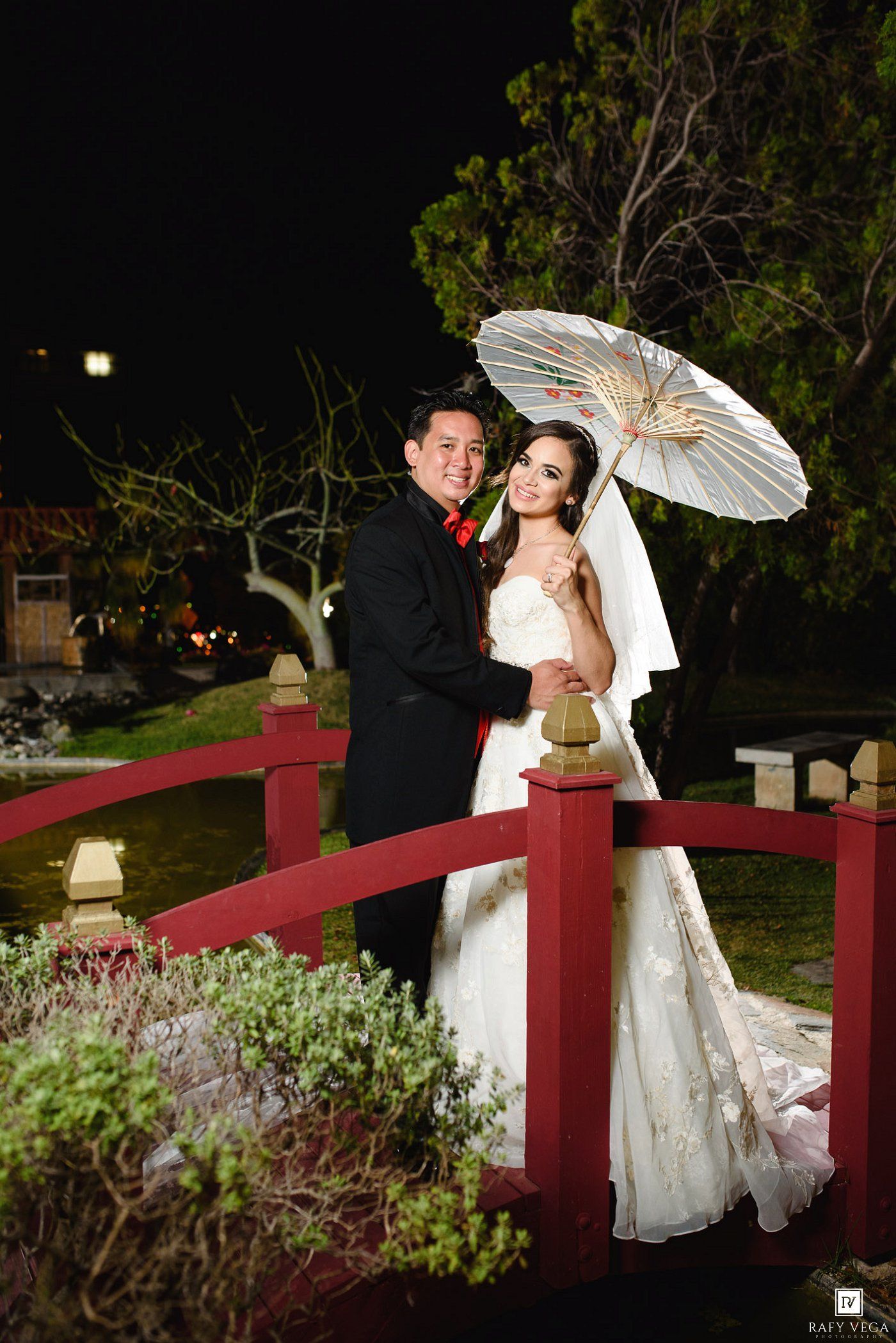 Bride and groom pose on red bridge at night, bride holding white parasol in elegant wedding gown.