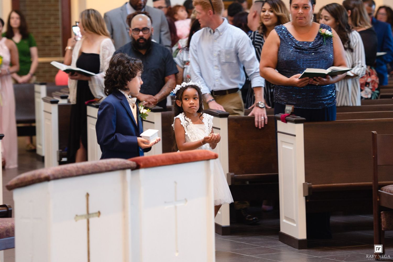 Two children in formal attire chat in church pews during a wedding ceremony with adults holding hymnals.