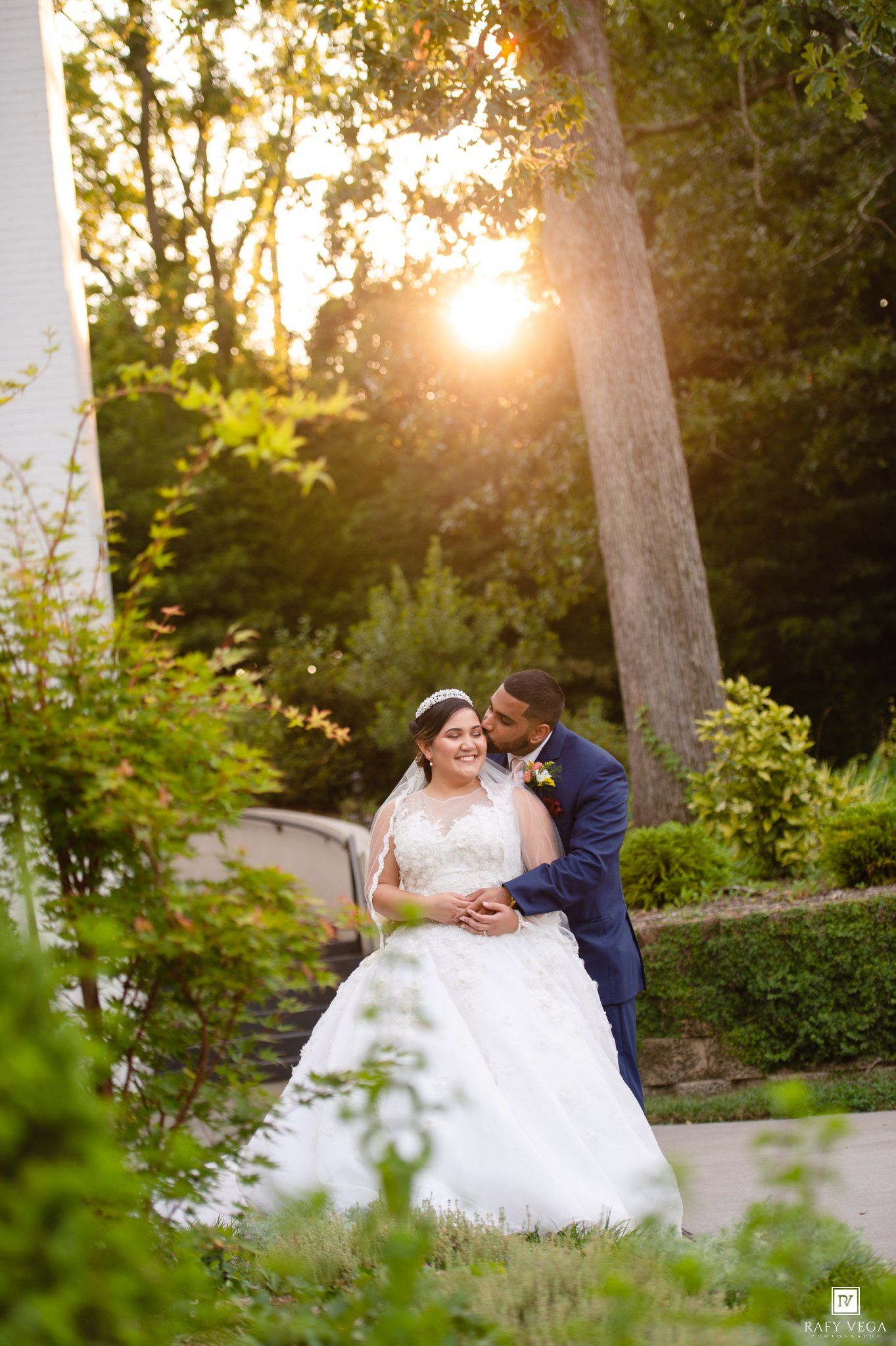 Bride and groom share a romantic moment in a lush garden at golden hour, with warm sunlight filtering through the trees.