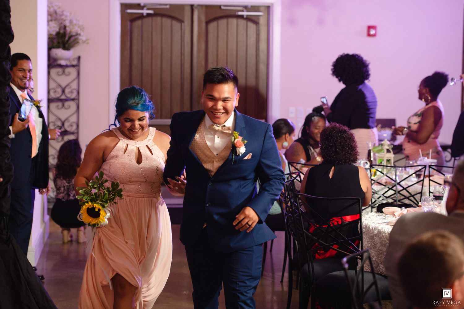 Bridesmaid in blush pink dress walks with groomsman in navy suit at wedding reception.
