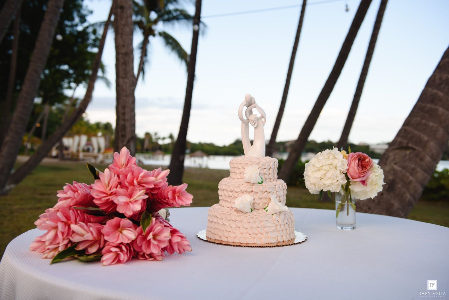 Three-tier white wedding cake with bride and groom topper on table with pink tropical flowers and white roses outdoors.