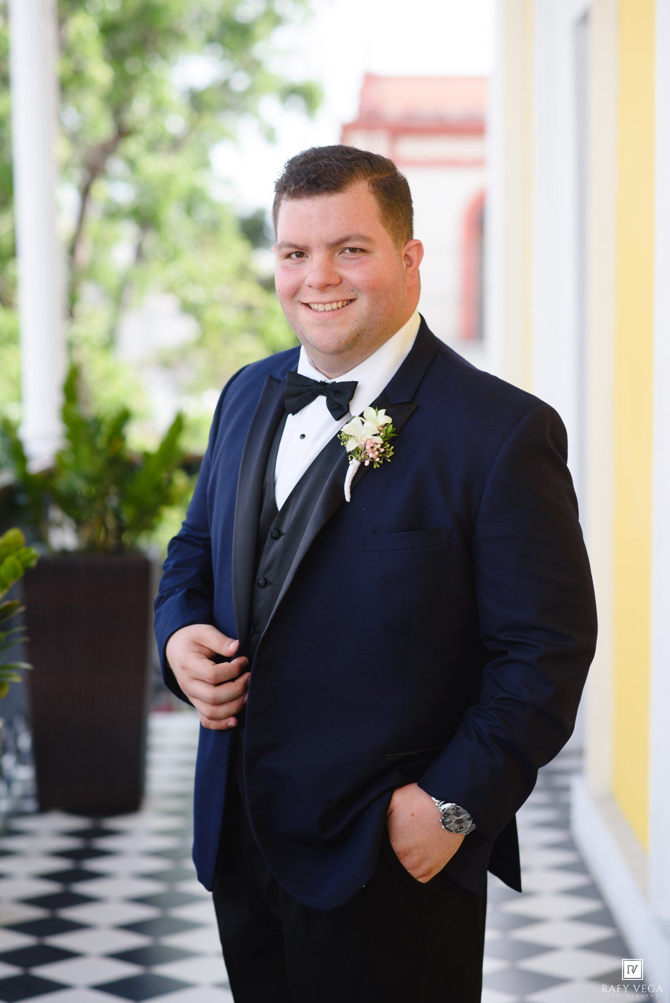 Smiling groom in navy tuxedo with bow tie and boutonniere poses on checkered floor porch.