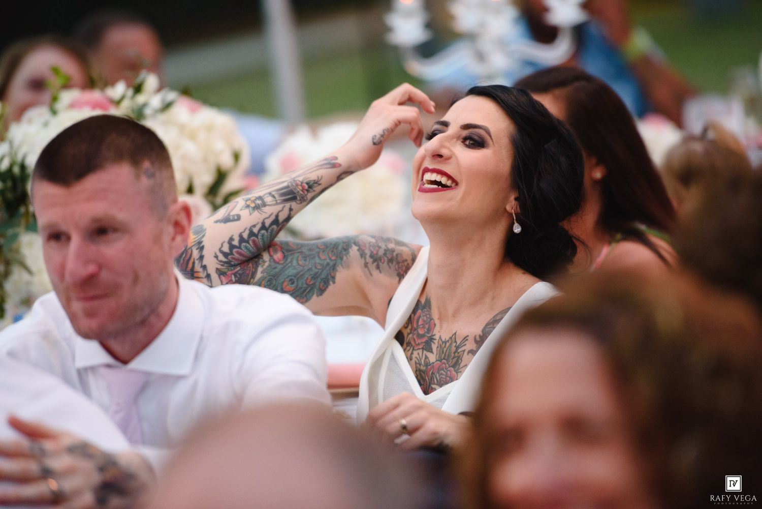 A tattooed bride laughs joyfully while seated at her outdoor wedding reception surrounded by guests.