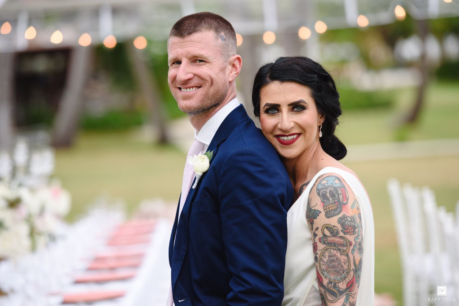 A smiling bride with a floral sleeve tattoo and groom in a navy suit pose back-to-back at an outdoor wedding reception.