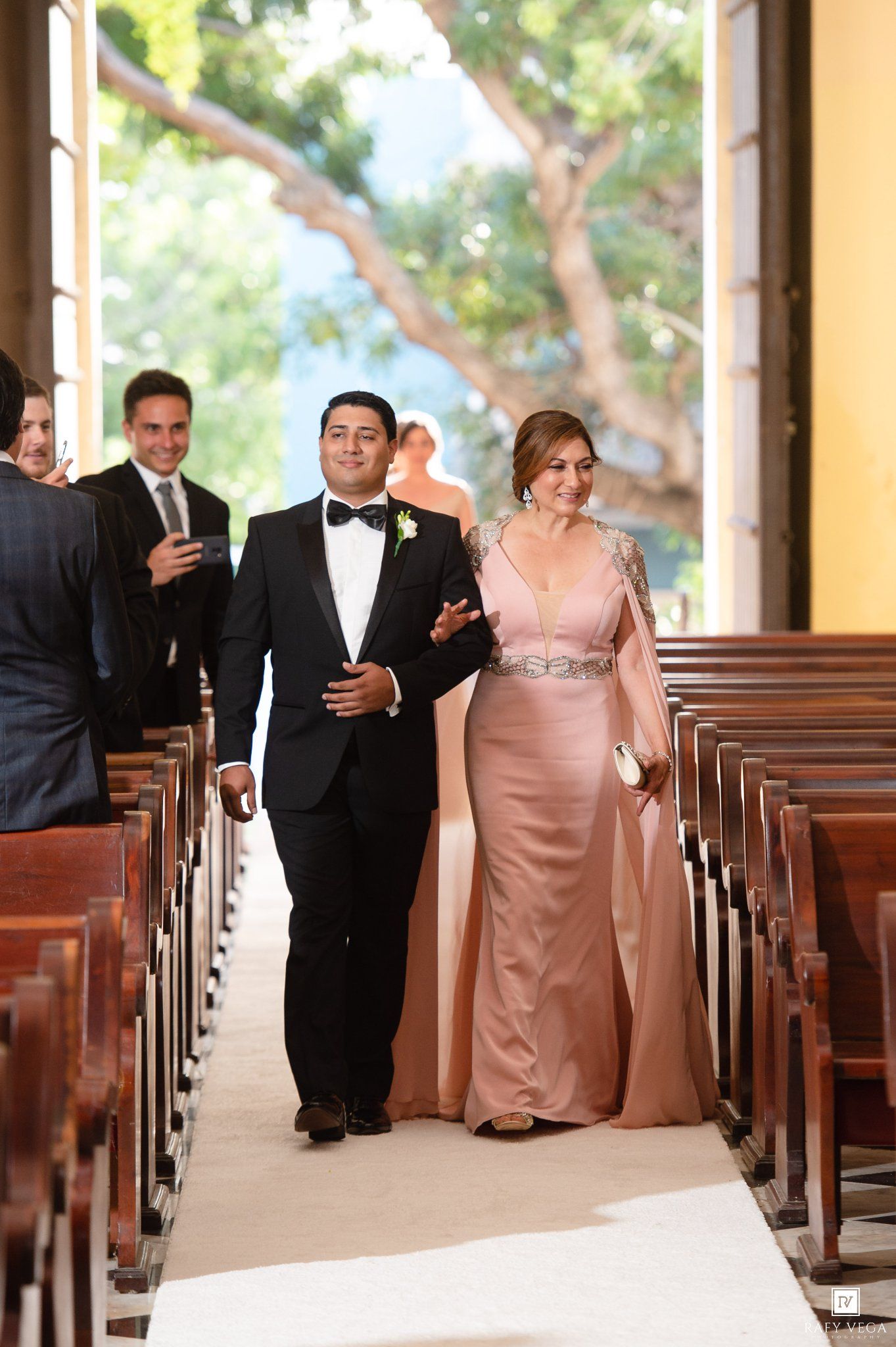 Groom in tuxedo walks down church aisle with elegant woman in pink gown during wedding ceremony.