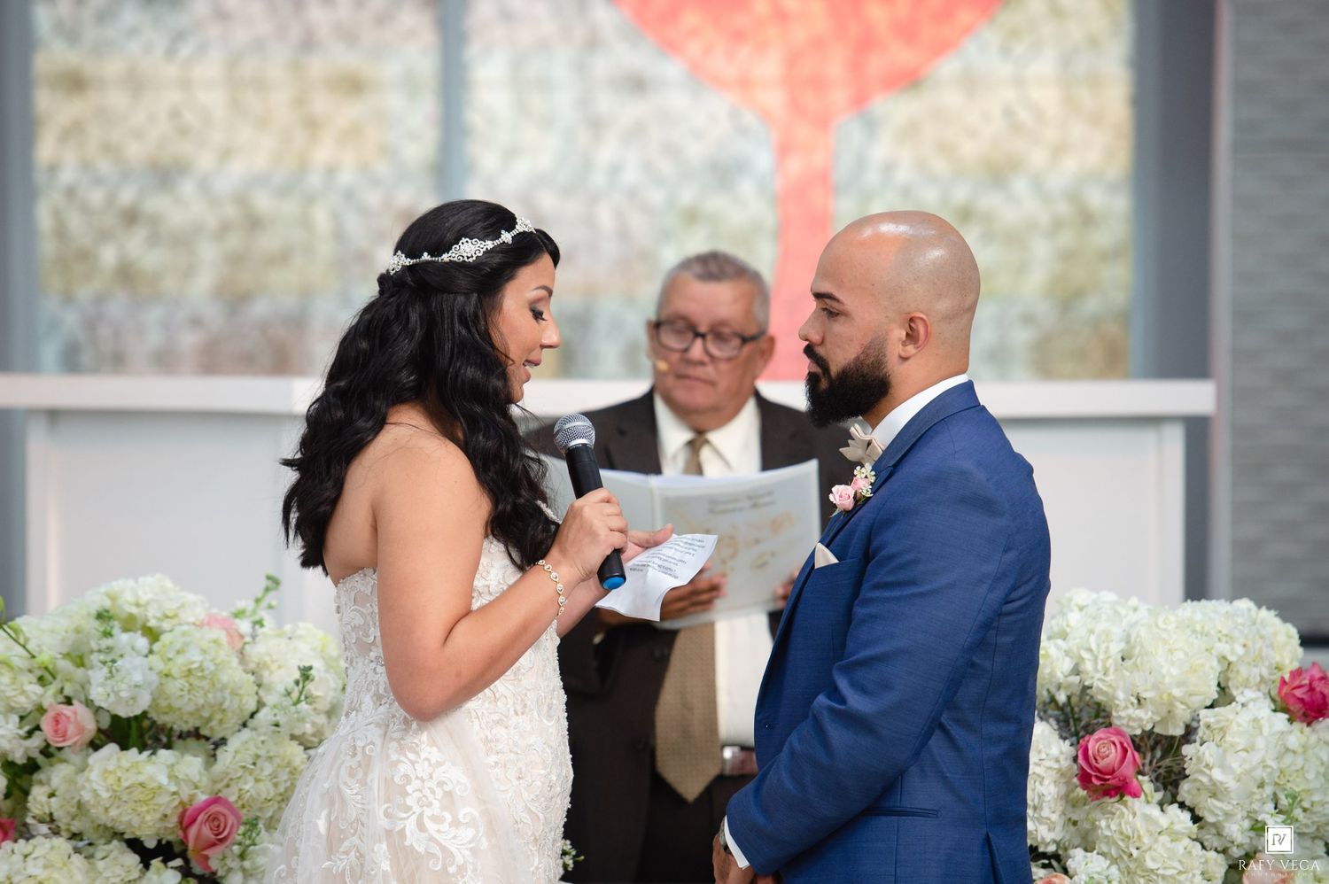 Bride reads wedding vows to groom at elegant ceremony with white floral arrangements and officiant in background.
