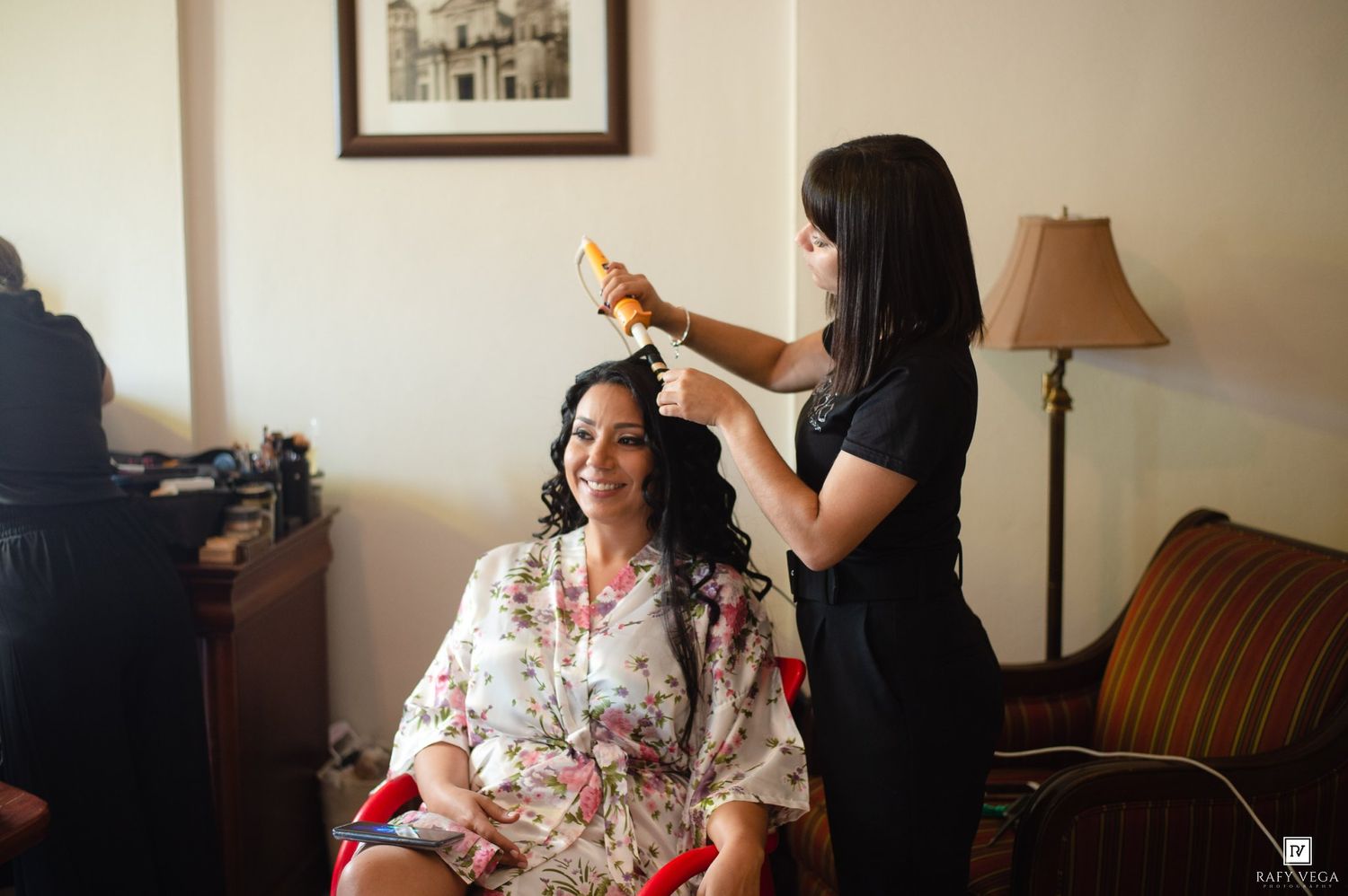 Bride in floral robe gets hair styled by professional hairstylist in bright hotel room before wedding.