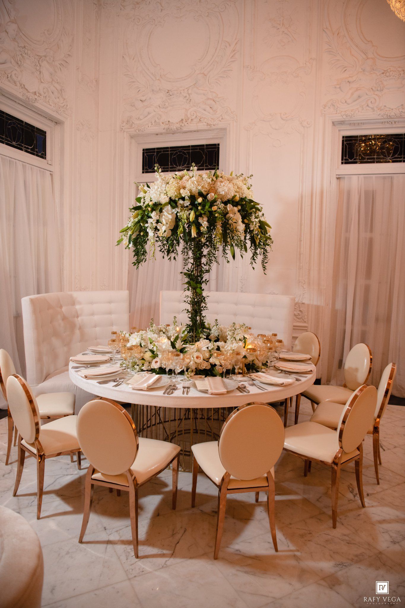 Elegant wedding reception table with tall floral centerpiece of white roses and greenery in ornate ballroom.