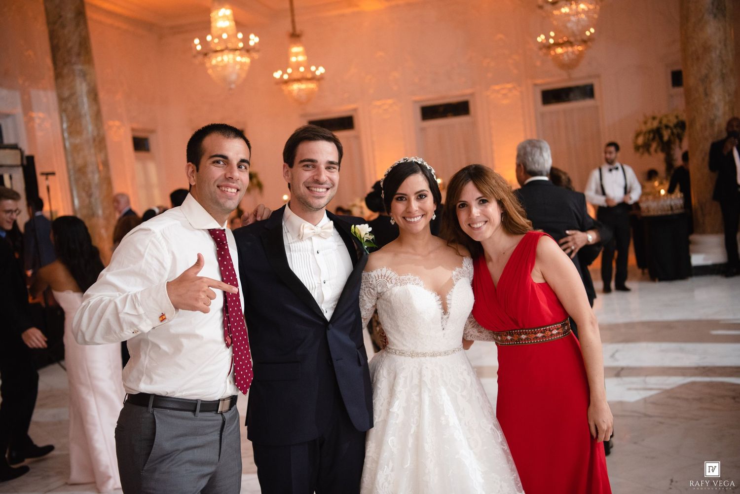 Bride in white gown and groom in tuxedo pose with two guests at elegant wedding reception venue.