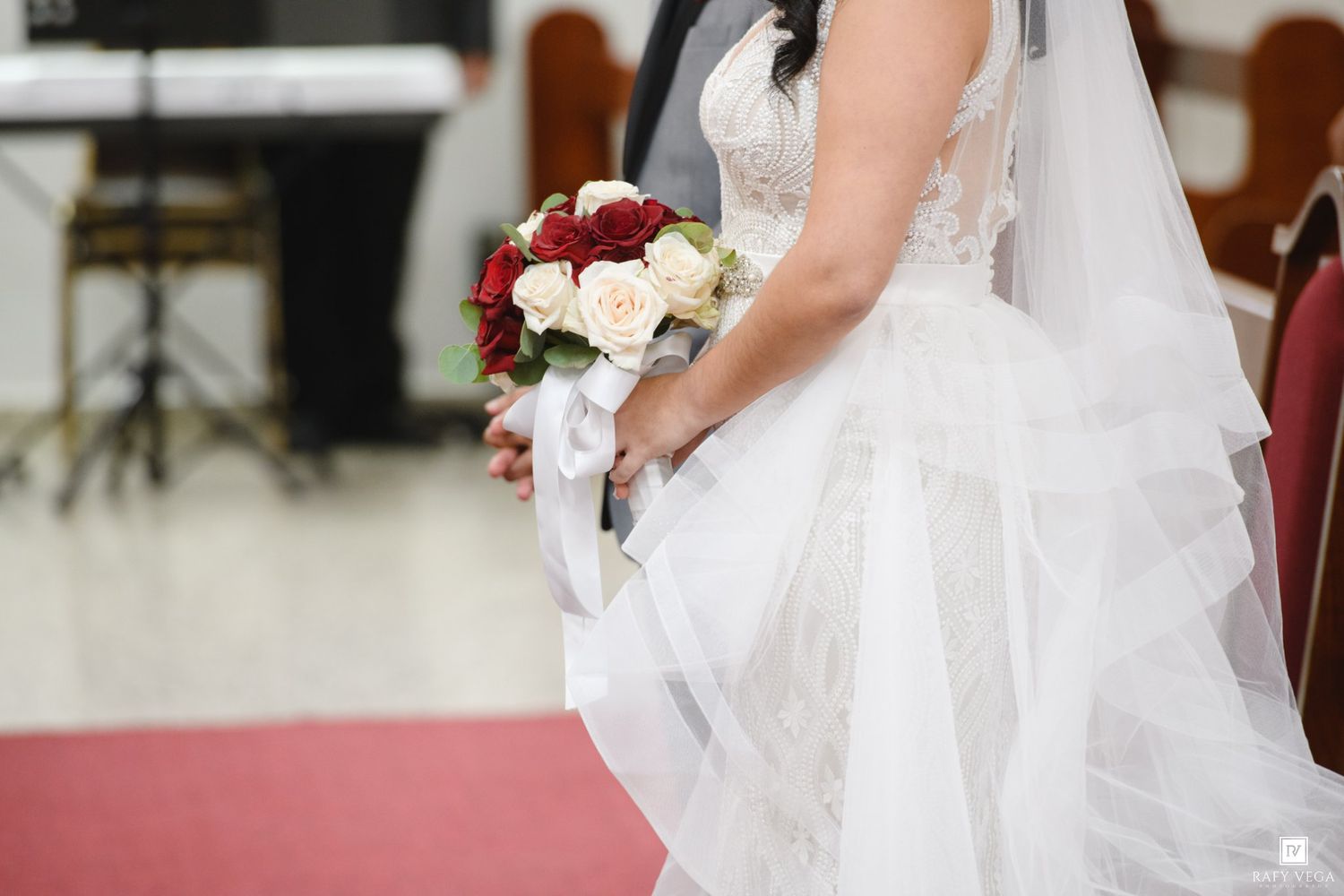 Bride in white lace wedding gown holding red and white rose bouquet at church ceremony.
