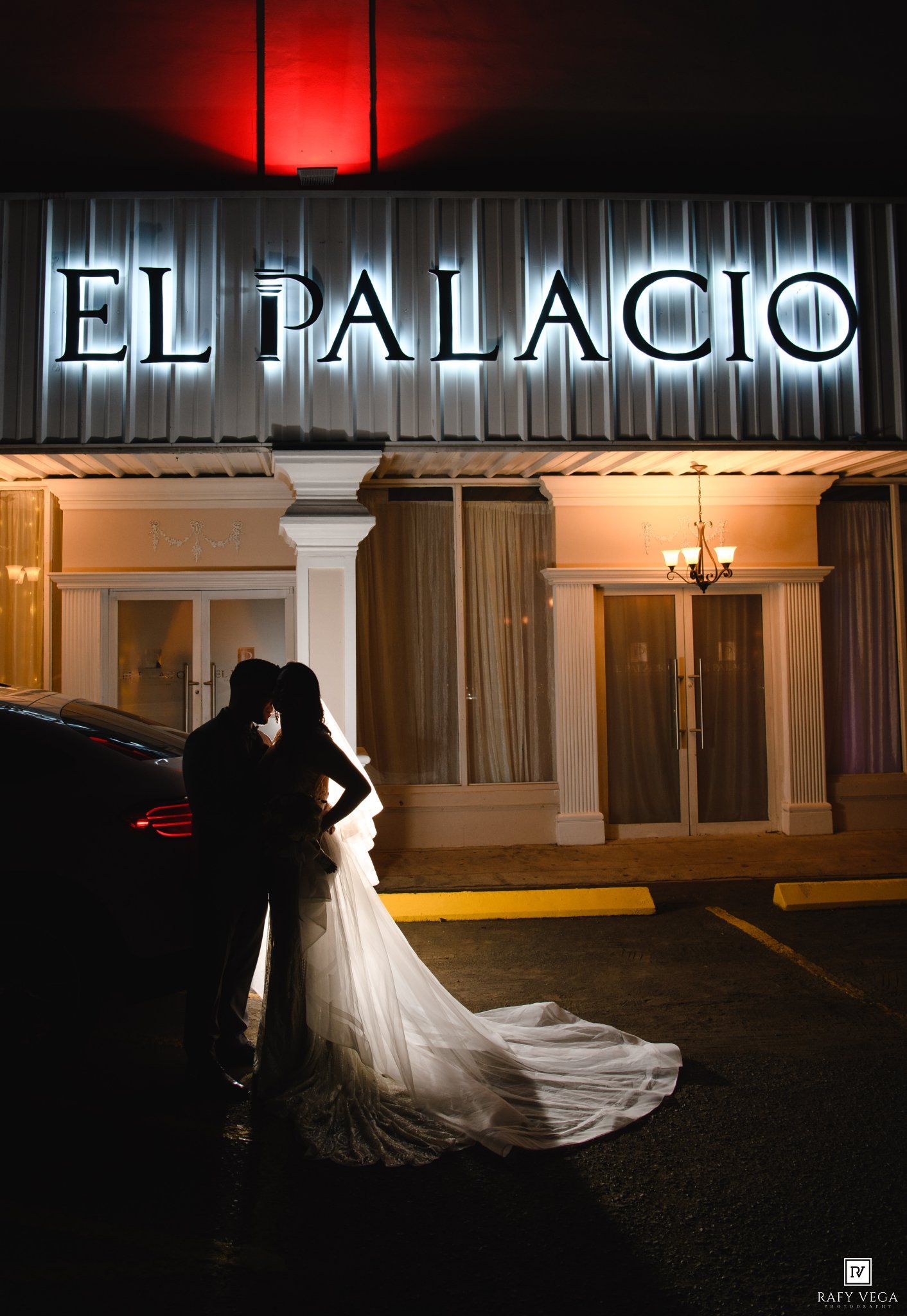 Silhouette of a bride and groom kissing outside El Palacio venue at night, with glowing sign and red accent lighting.