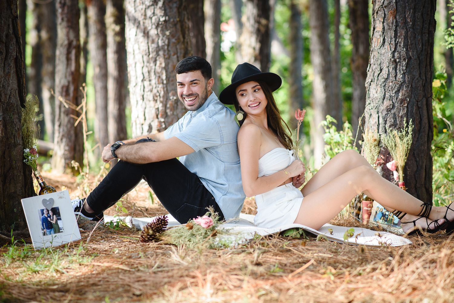 Couple sitting back-to-back smiling at a forest picnic, woman in white dress and black hat, man in light blue shirt.