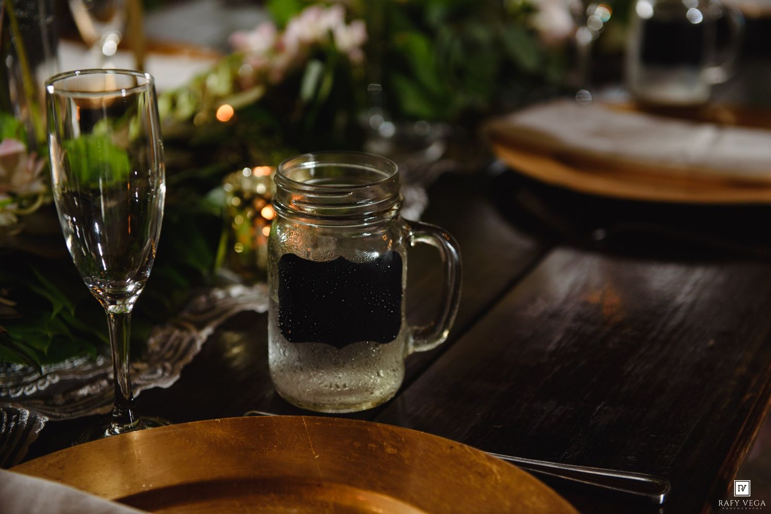 Mason jar drink with chalkboard label on elegant gold plate wedding table setting with champagne flute and greenery.