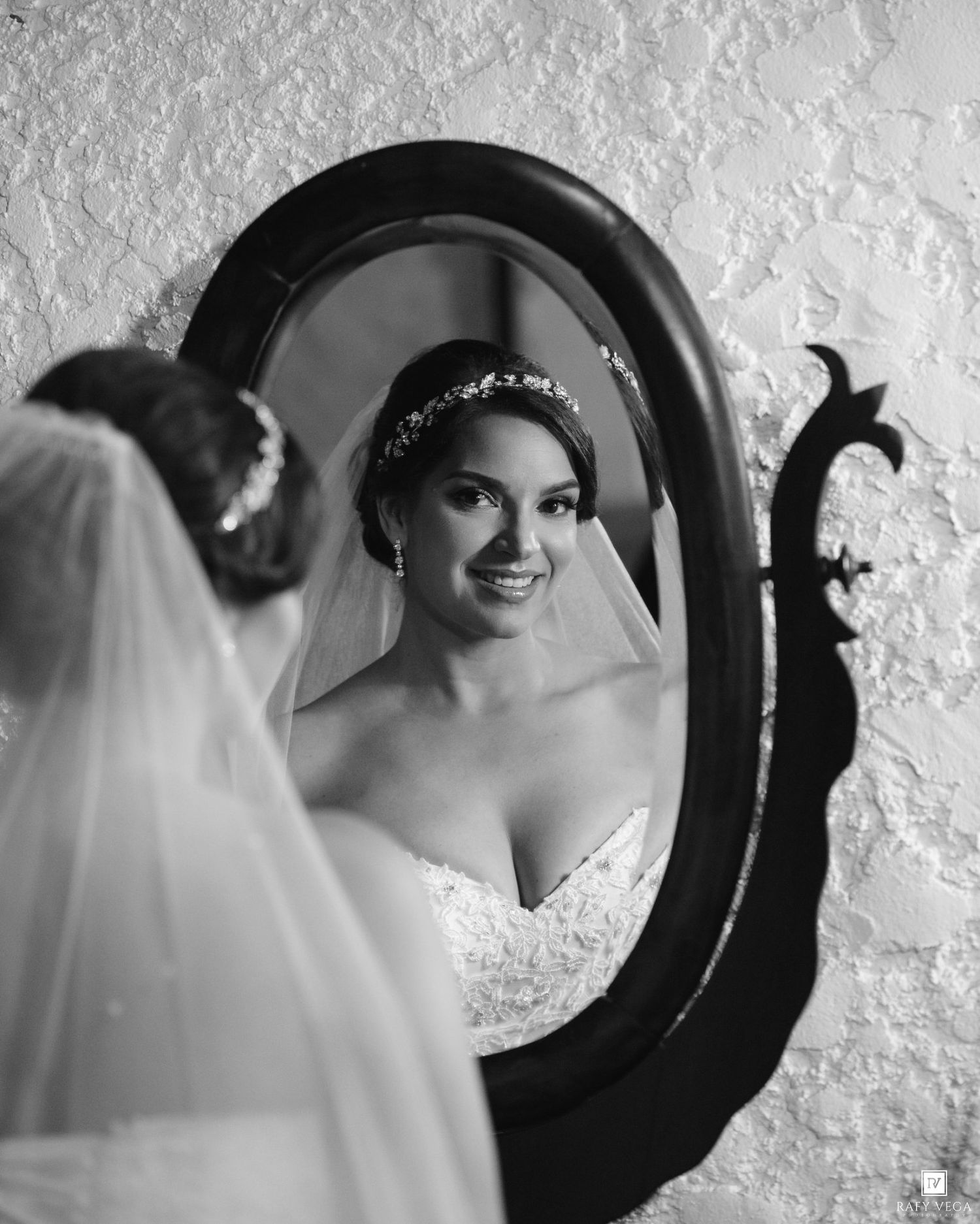 Bride smiling in ornate oval mirror reflection, wearing lace wedding dress and floral headpiece with veil, black and white photo.