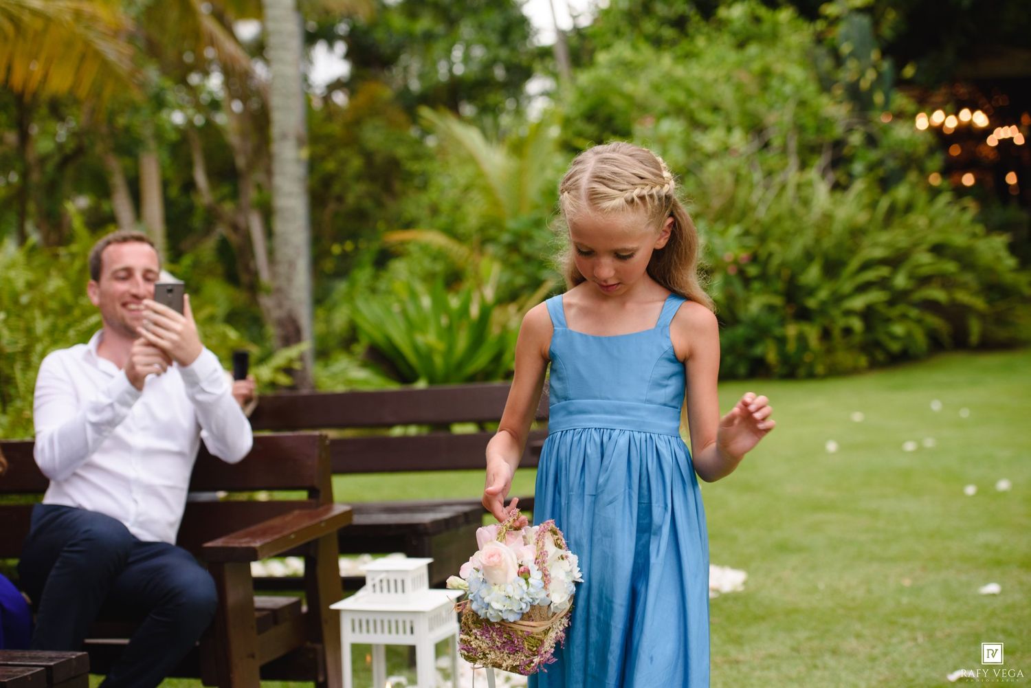 Young flower girl in blue dress scattering petals at outdoor wedding ceremony with guest photographing.